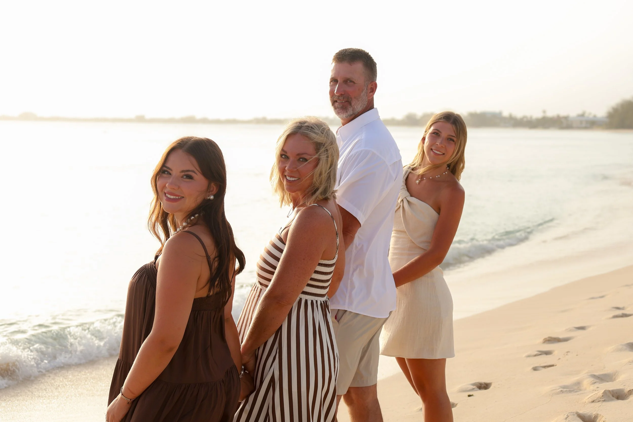 A group of five people, four women and one man, standing in a line on a beach during sunset, smiling and facing the camera, with the ocean and distant shoreline in the background.