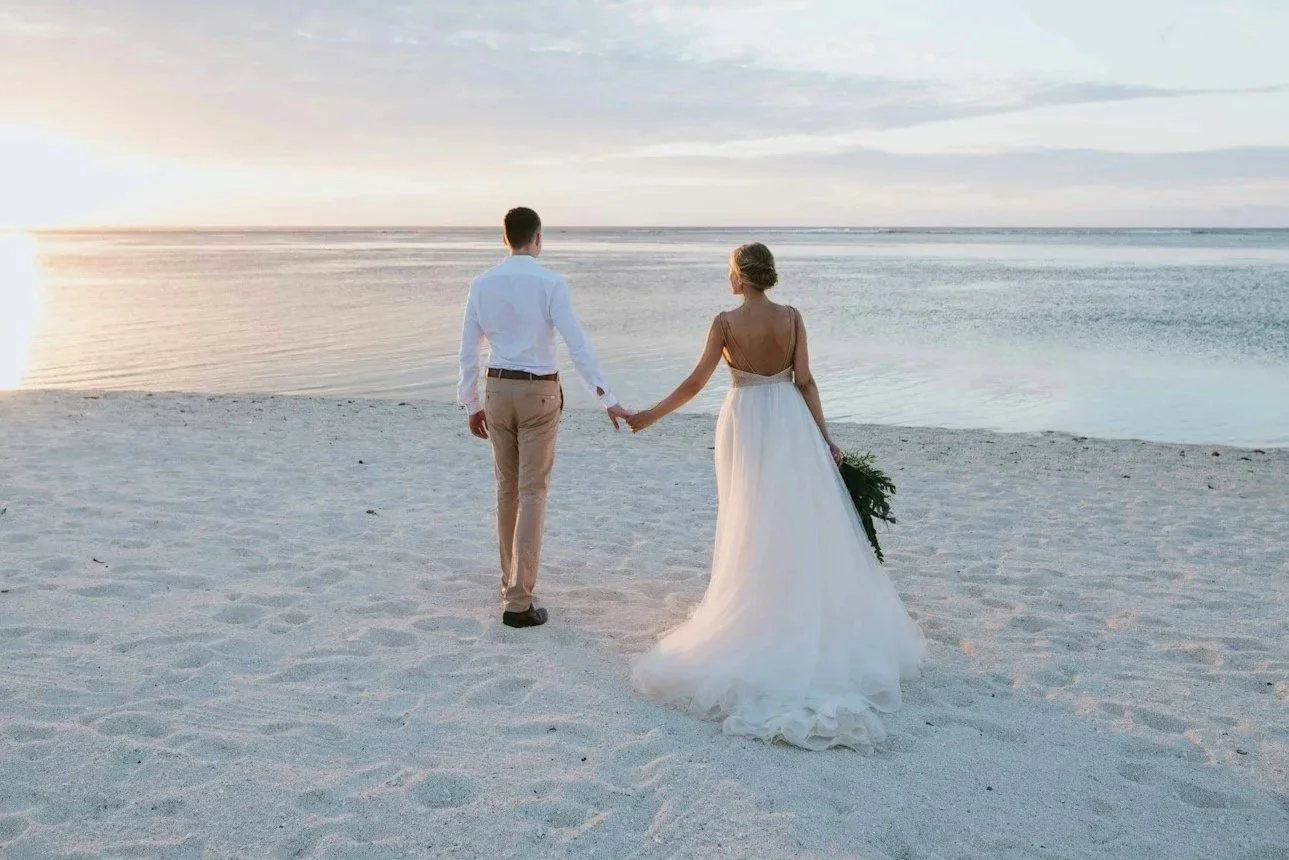 A bride and groom holding hands on a beach at sunset.