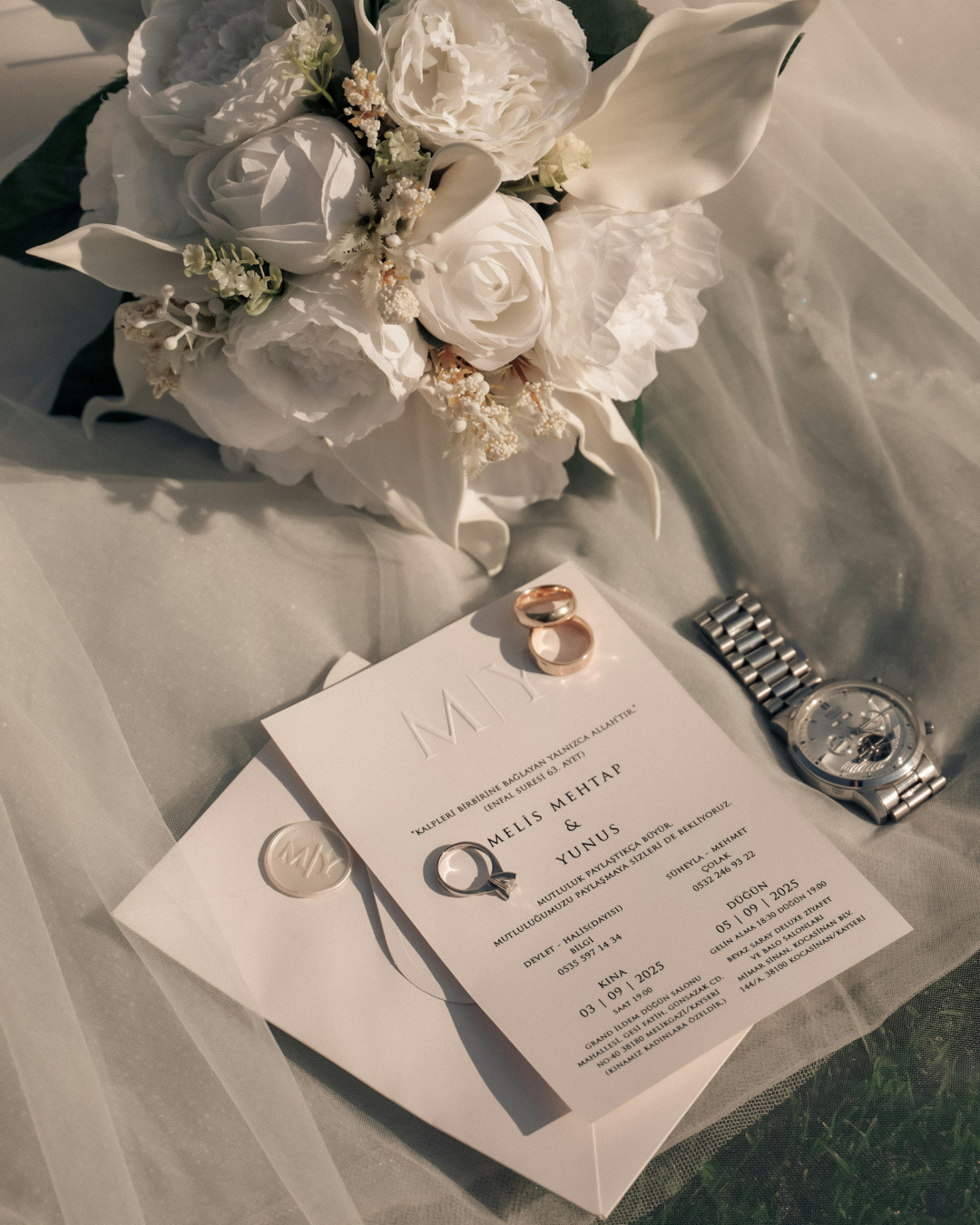Wedding bouquet of white roses and calla lilies, wedding rings, a wristwatch, and an invitation card on a table.