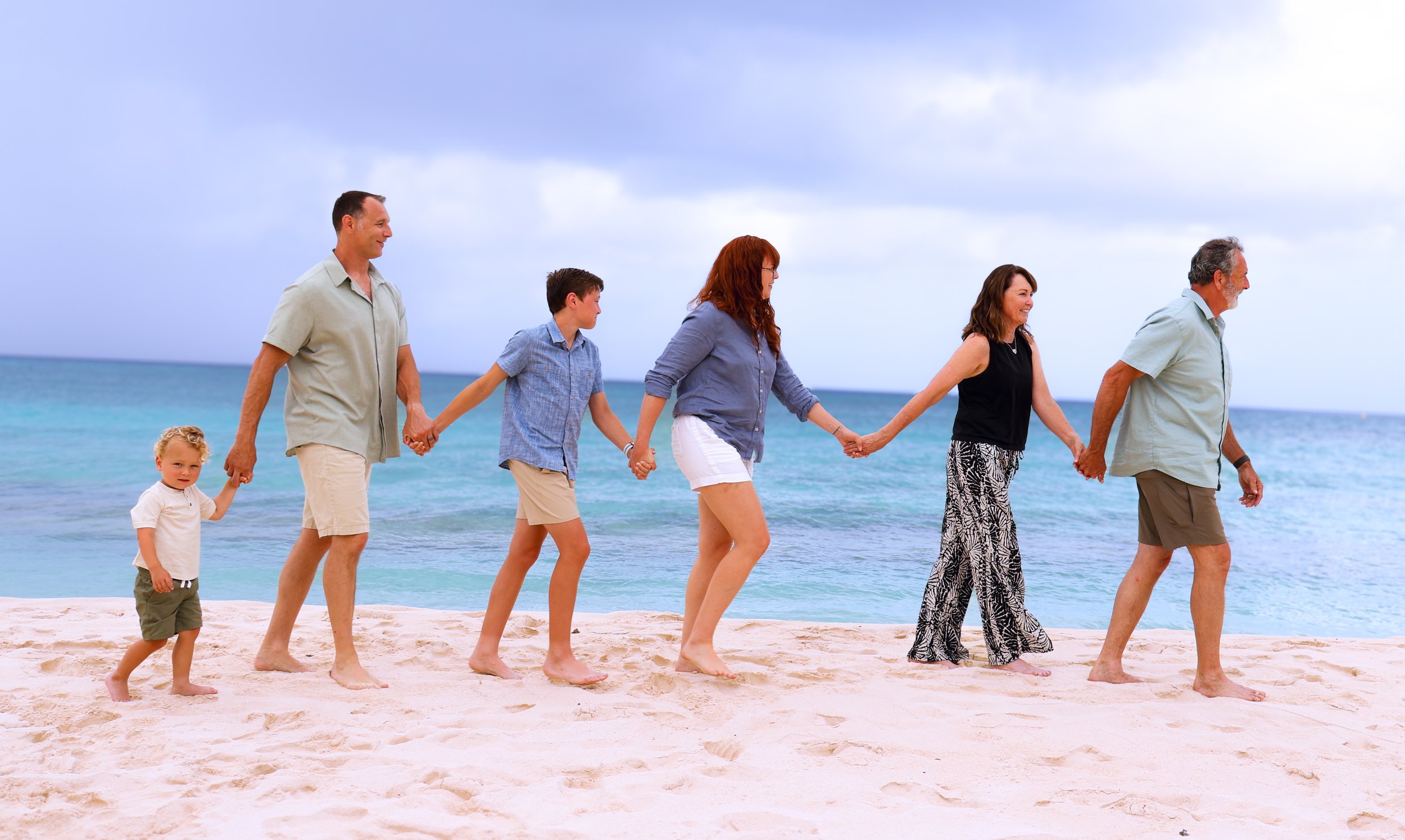 A family walking on the beach holding hands, with the ocean in the background. The group includes a child, two men, two women, all smiling and enjoying the seaside.