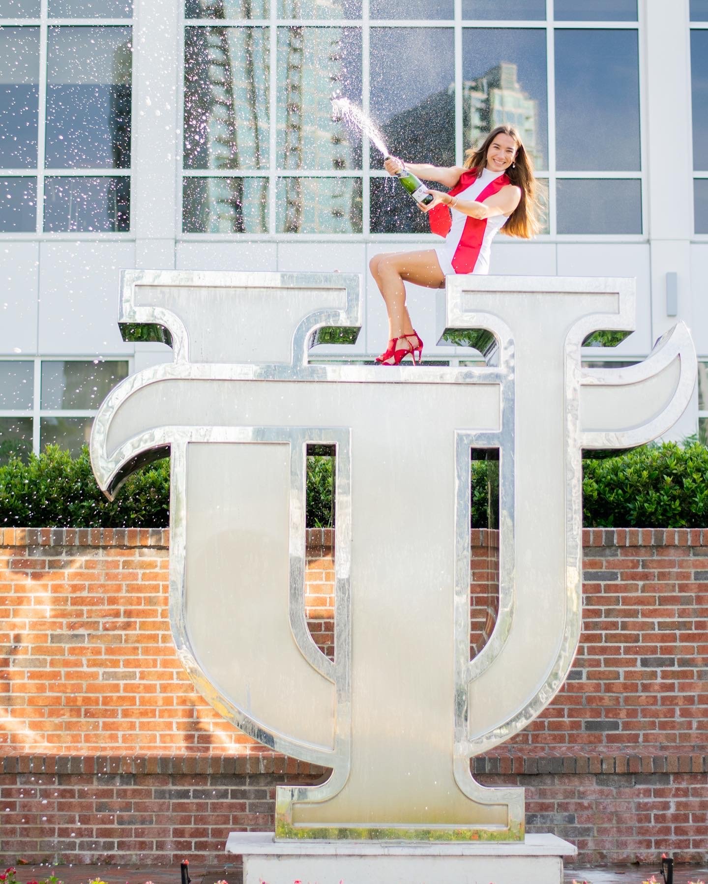 A woman in a white and red dress and red high heels celebrating by spraying champagne while sitting on a large metallic Greek letter sigma outside a modern building with glass windows.