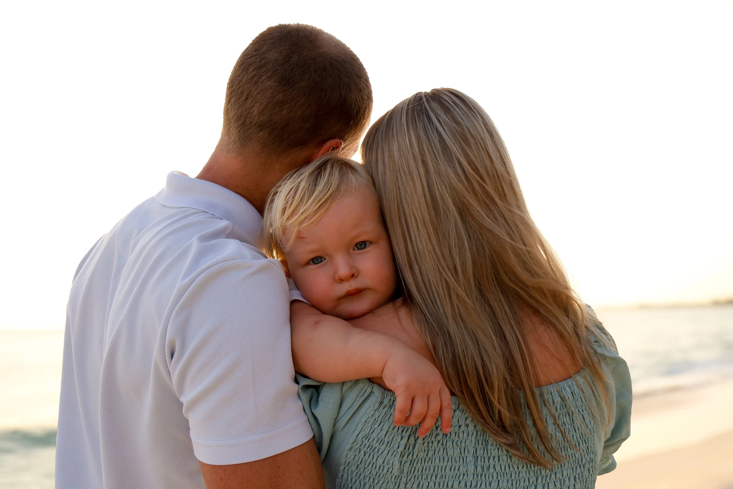 A family of three hugging on the beach during sunset, with the ocean in the background.