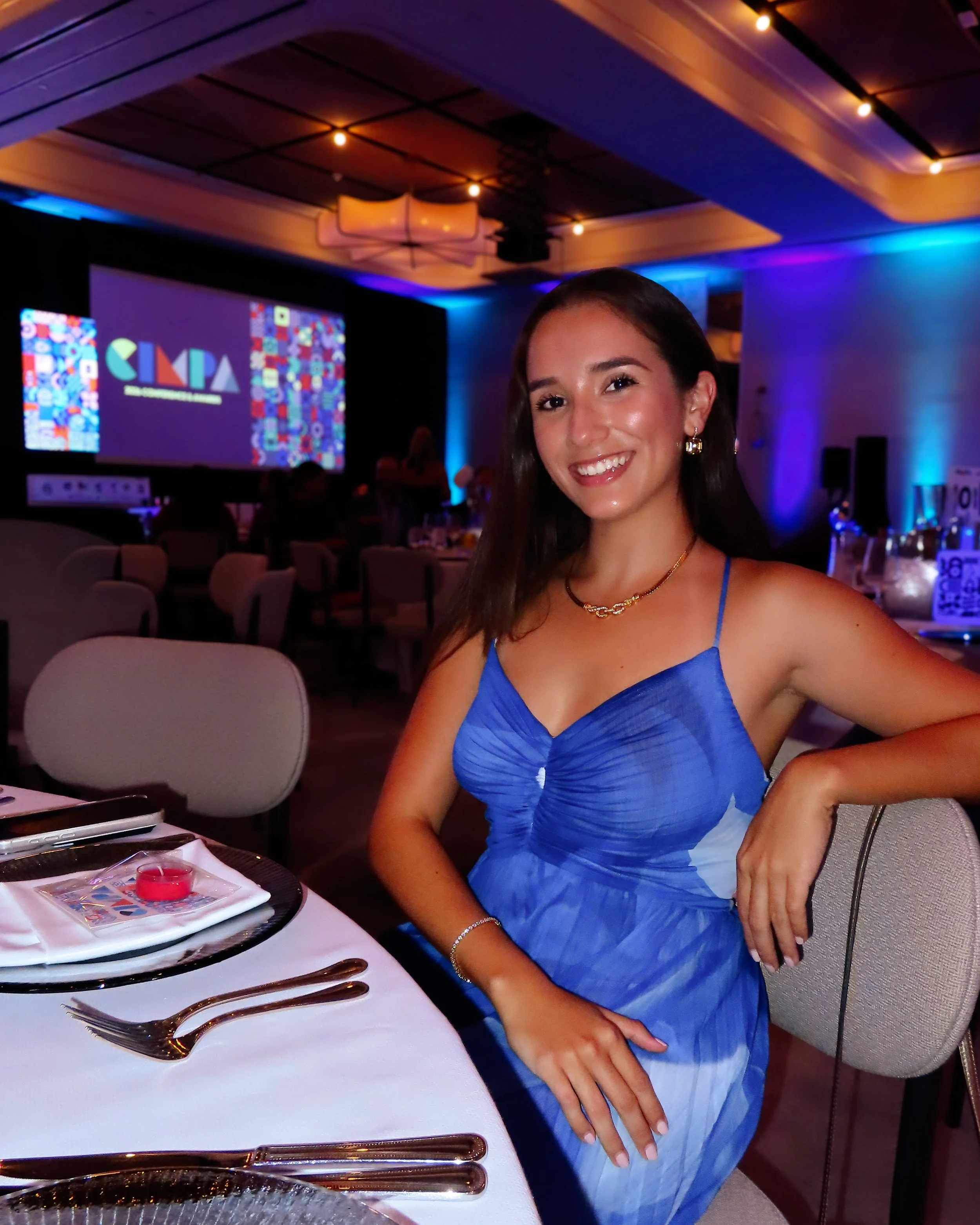 A young woman in a blue dress sitting at a table set for a formal dinner, smiling at the camera, with a decorated banquet hall in the background.