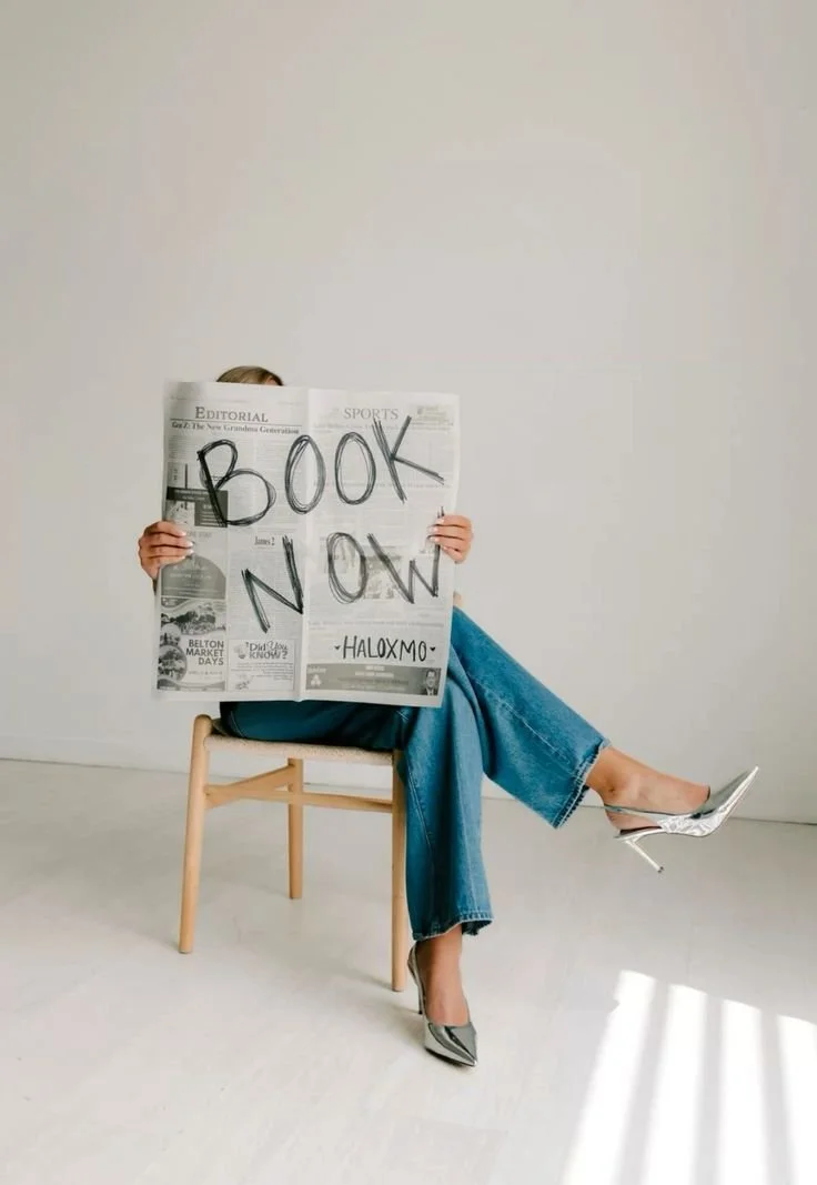 Person sitting on a wooden chair, holding a newspaper with 'BOOK NOW' written on it, wearing high heels, jeans, and a blue top, against a plain white wall.