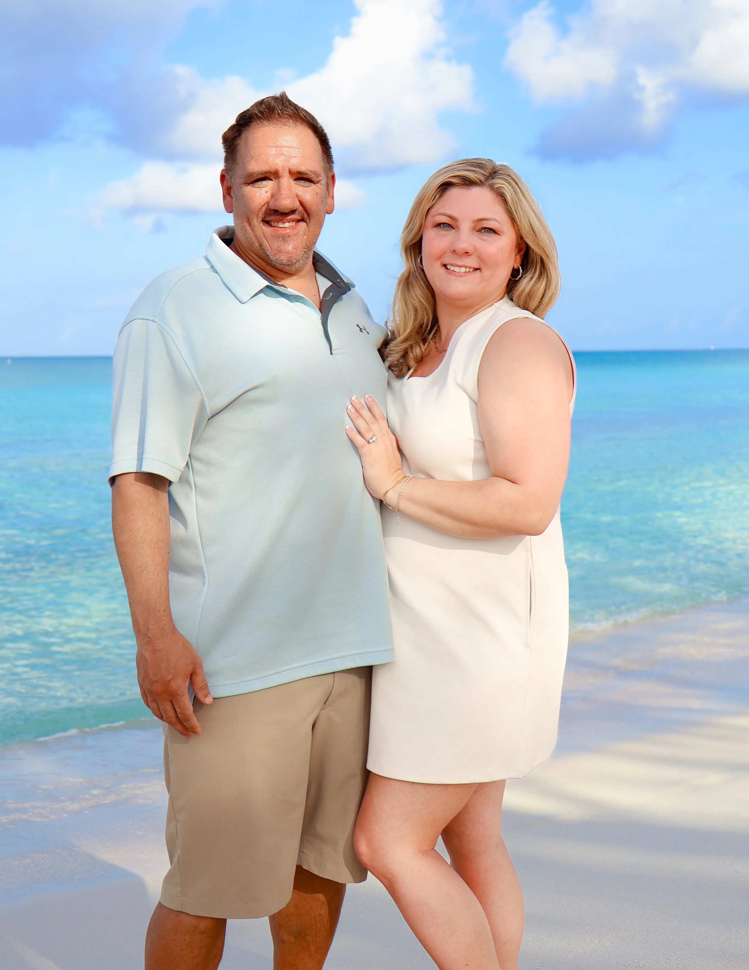 A man and woman standing on a beach with clear blue water and partly cloudy sky in the background. The man is wearing a light gray polo shirt and khaki shorts. The woman is wearing a white sleeveless dress and has blonde hair.