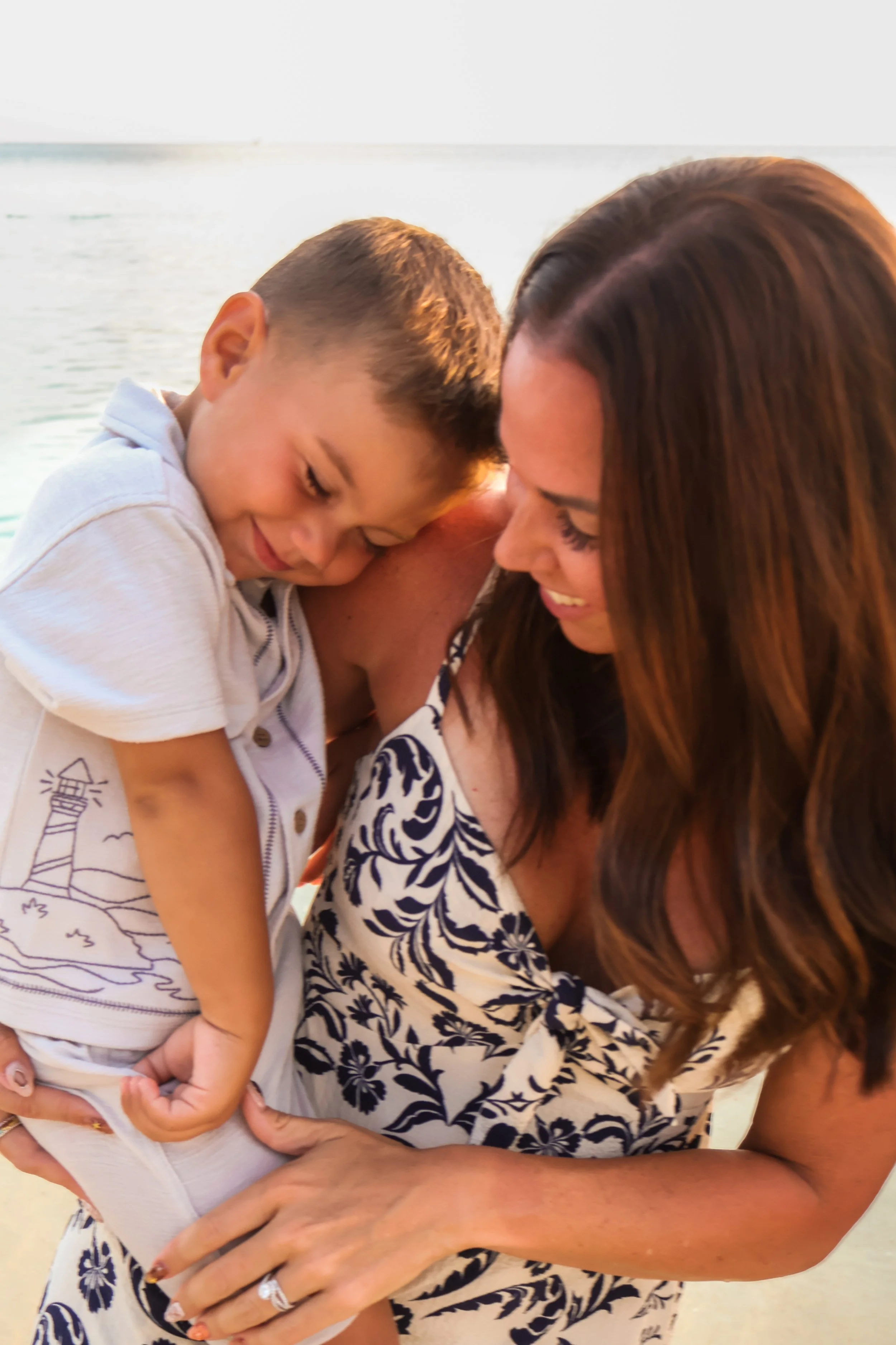 A woman with long brown hair holding a young boy with short hair in her arms at the beach. Both have their eyes closed and are smiling.
