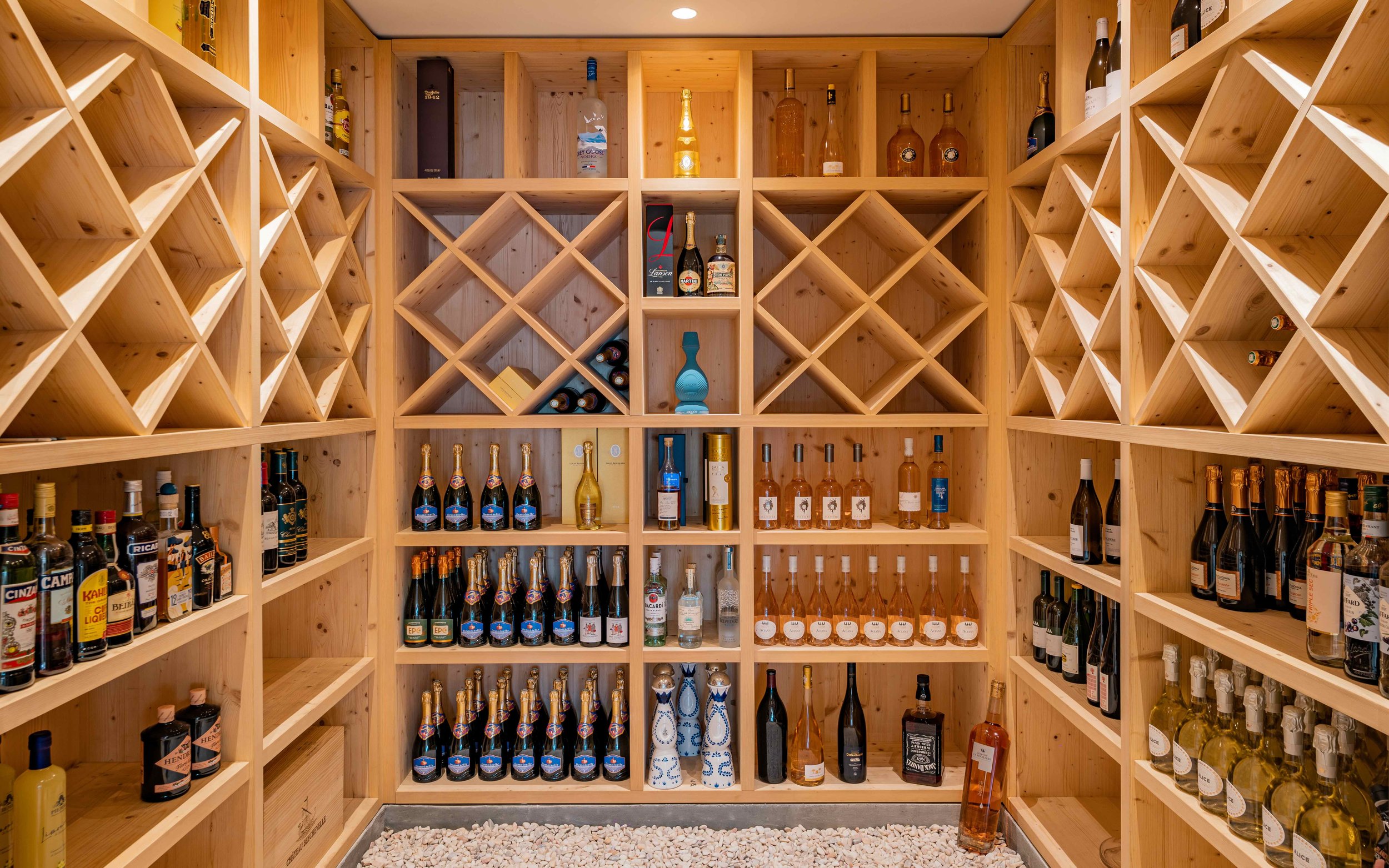 Wooden wine cellar with shelves filled with various bottles of wine and spirits, including champagne and liquor, and a gravel floor.