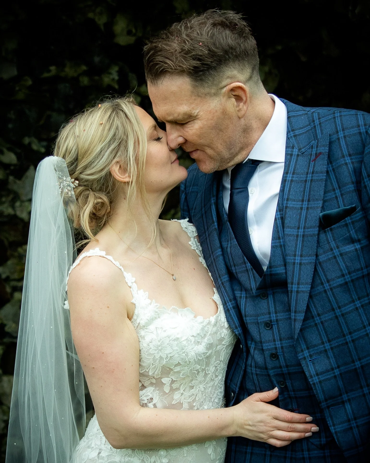A bride and groom in a close, intimate pose, about to kiss, outdoors with dark foliage in the background.