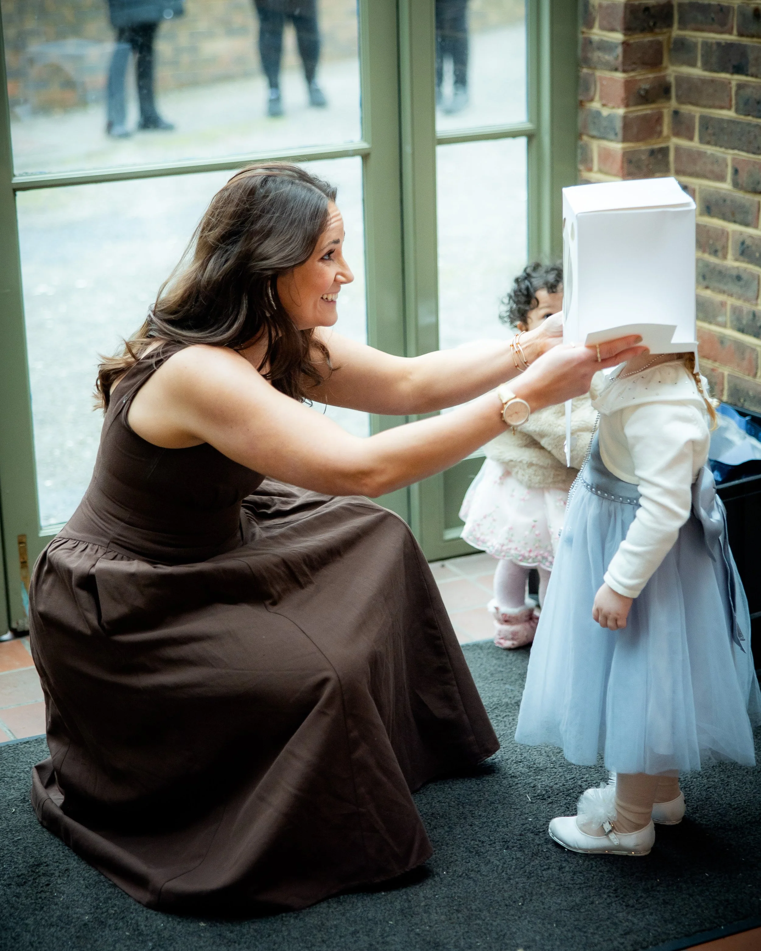 Woman smiling and playfully lifting a child's head with a box in an indoor setting with large windows and a brick wall.