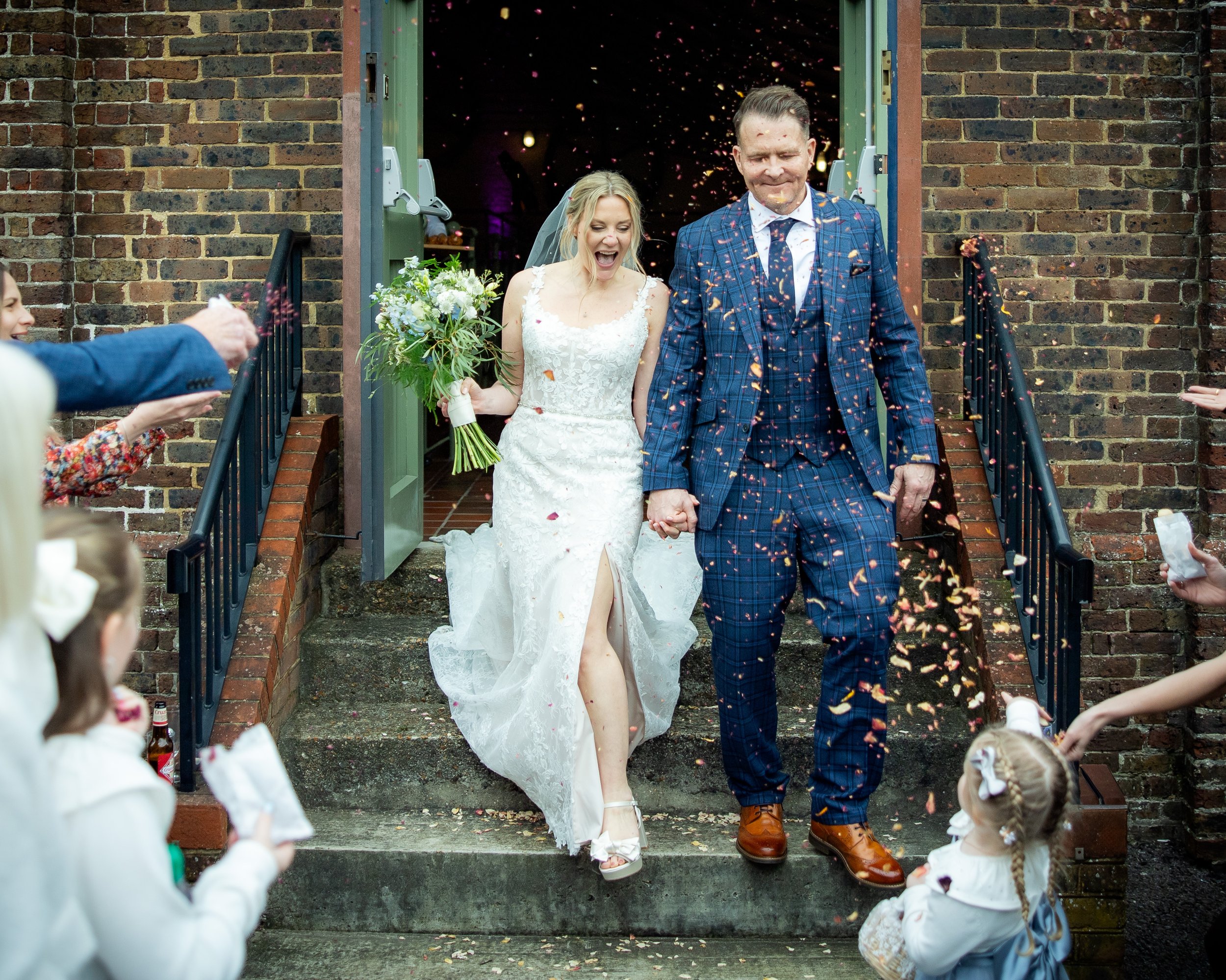 A newlywed couple walking out of a brick building on stairs, holding hands, as guests throw confetti and celebrate. The bride is in a white lace wedding dress, holding a bouquet. The groom is in a blue plaid suit. Children and adults surround them, s