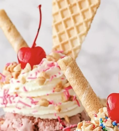 Close-up of a colorful ice cream sundae topped with a cherry, sprinkles, and crushed nuts, with waffle cookie and rolled wafer.