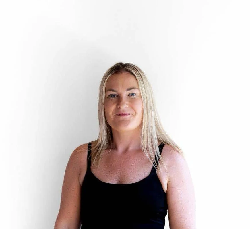 A woman with blonde hair wearing a black tank top, standing against a white background, smiling softly.