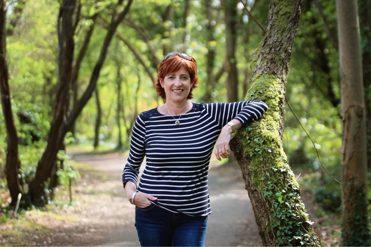 A woman with short red hair and a striped navy and white shirt standing outdoors in a wooded area, leaning on a tree trunk.