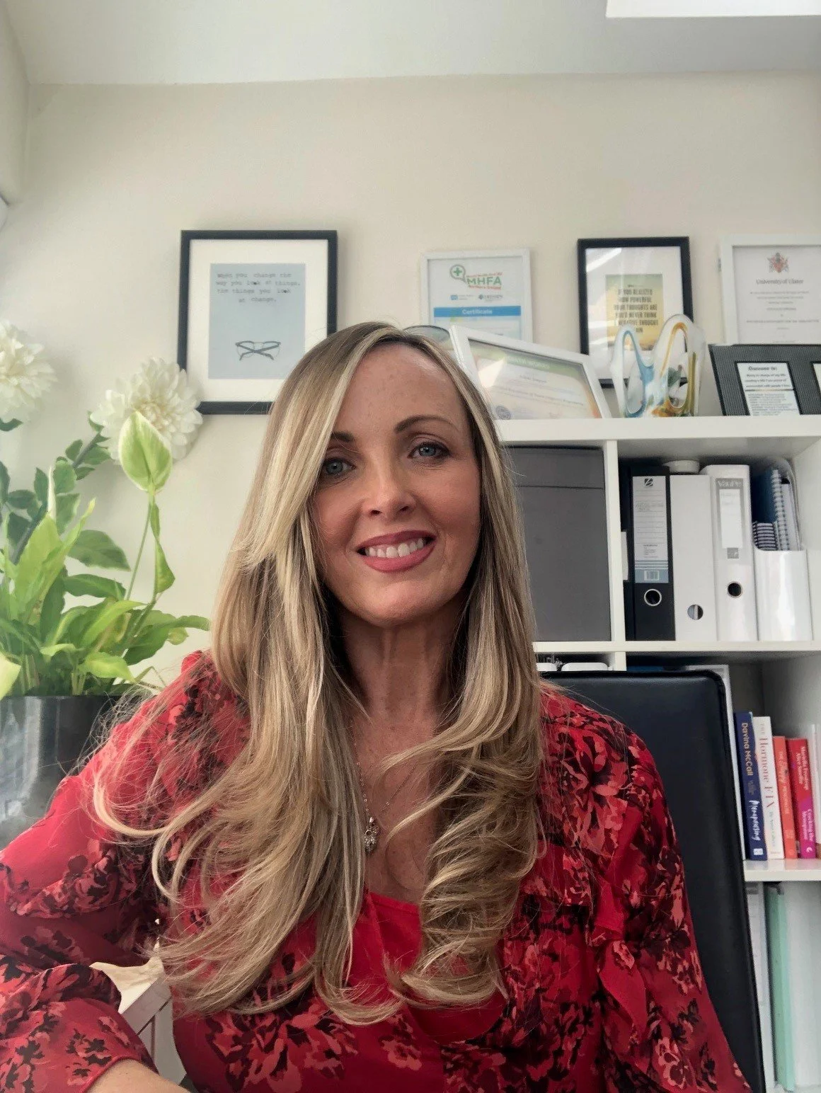 A woman with long blonde hair sitting in an office, wearing a red patterned top, smiling, with framed certificates and documents on the wall behind her, and a white bookshelf with binders and books.