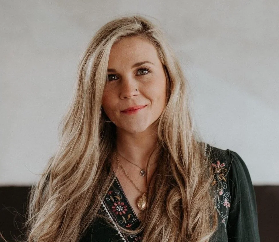 A young woman with long blonde hair, wearing a black embroidered top and layered necklaces, smiling softly at the camera.