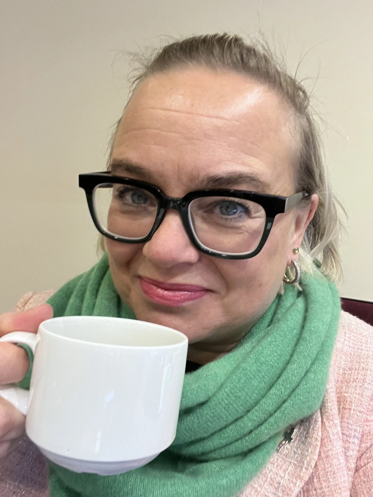A woman with glasses and earrings holding a white mug, smiling, wearing a green scarf and a pink jacket.
