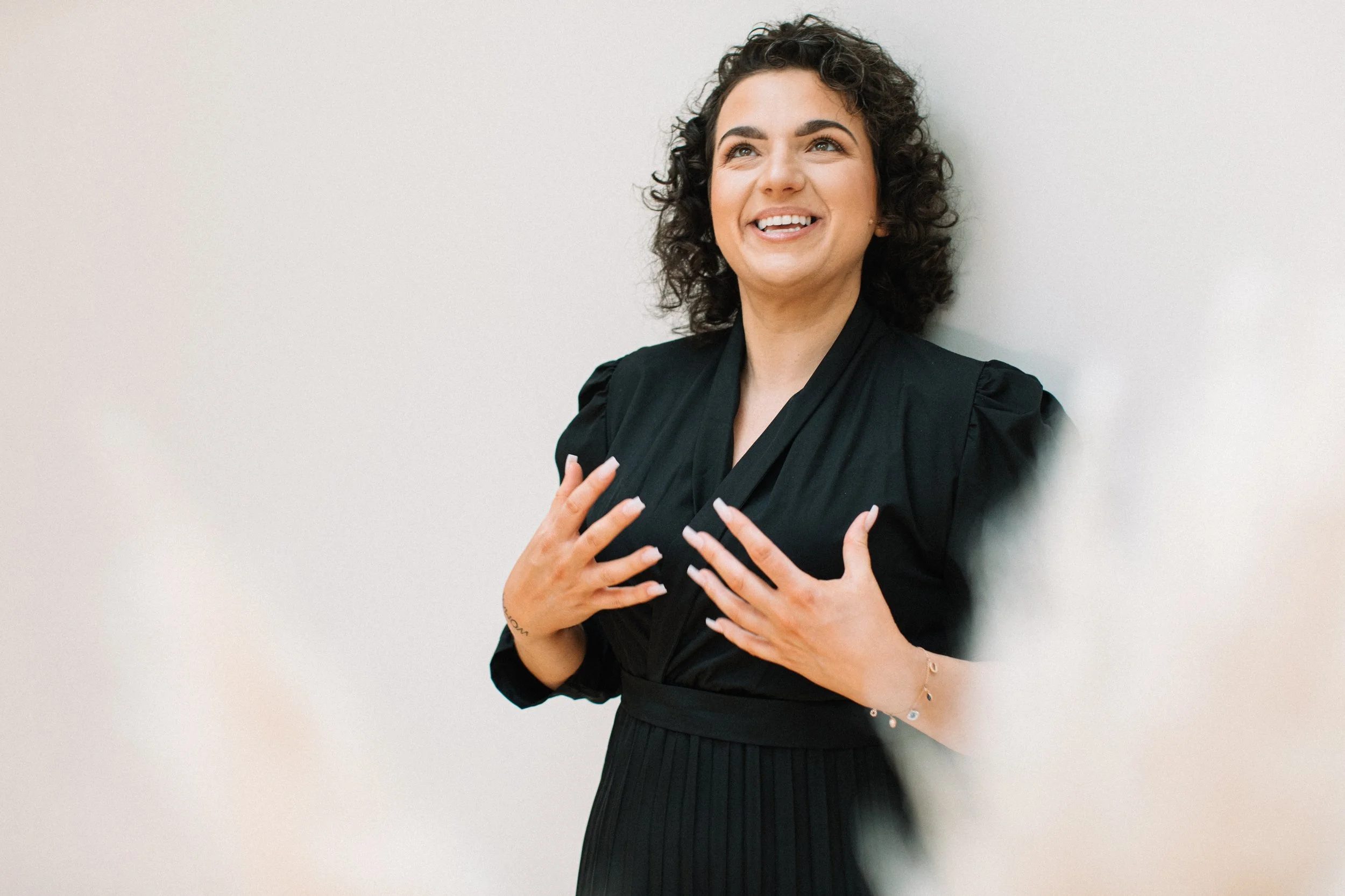 A woman with curly dark hair and light skin, smiling and wearing a black dress, standing against a plain light wall.