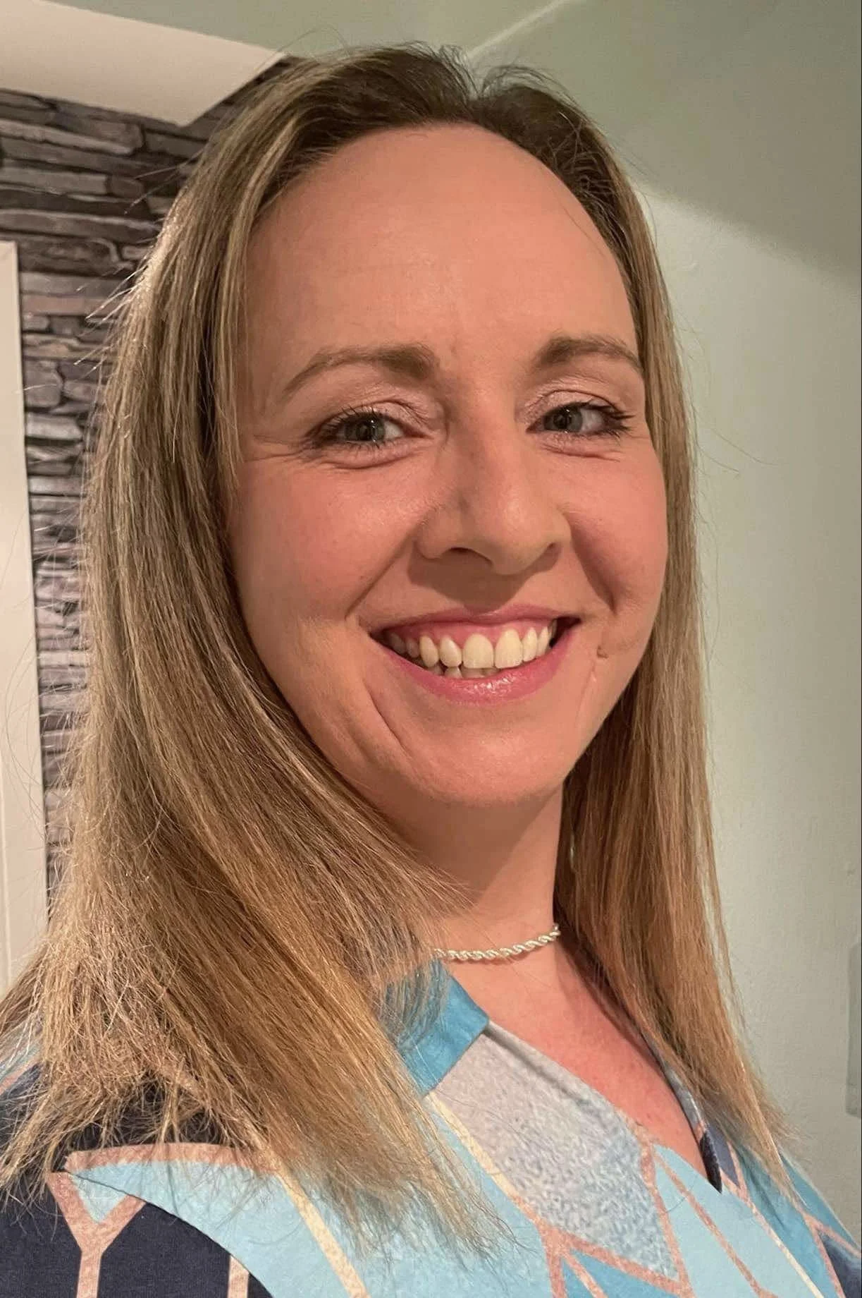 A woman smiling with long, light brown hair, wearing a colorful top with geometric patterns, a pearl necklace, and makeup, standing indoors with a stone accent wall and light green walls in the background.