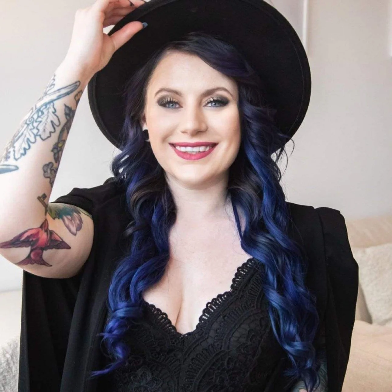 A woman smiling, wearing a black hat and black clothing, with long wavy hair dyed blue, and tattoos on her left arm, indoors.