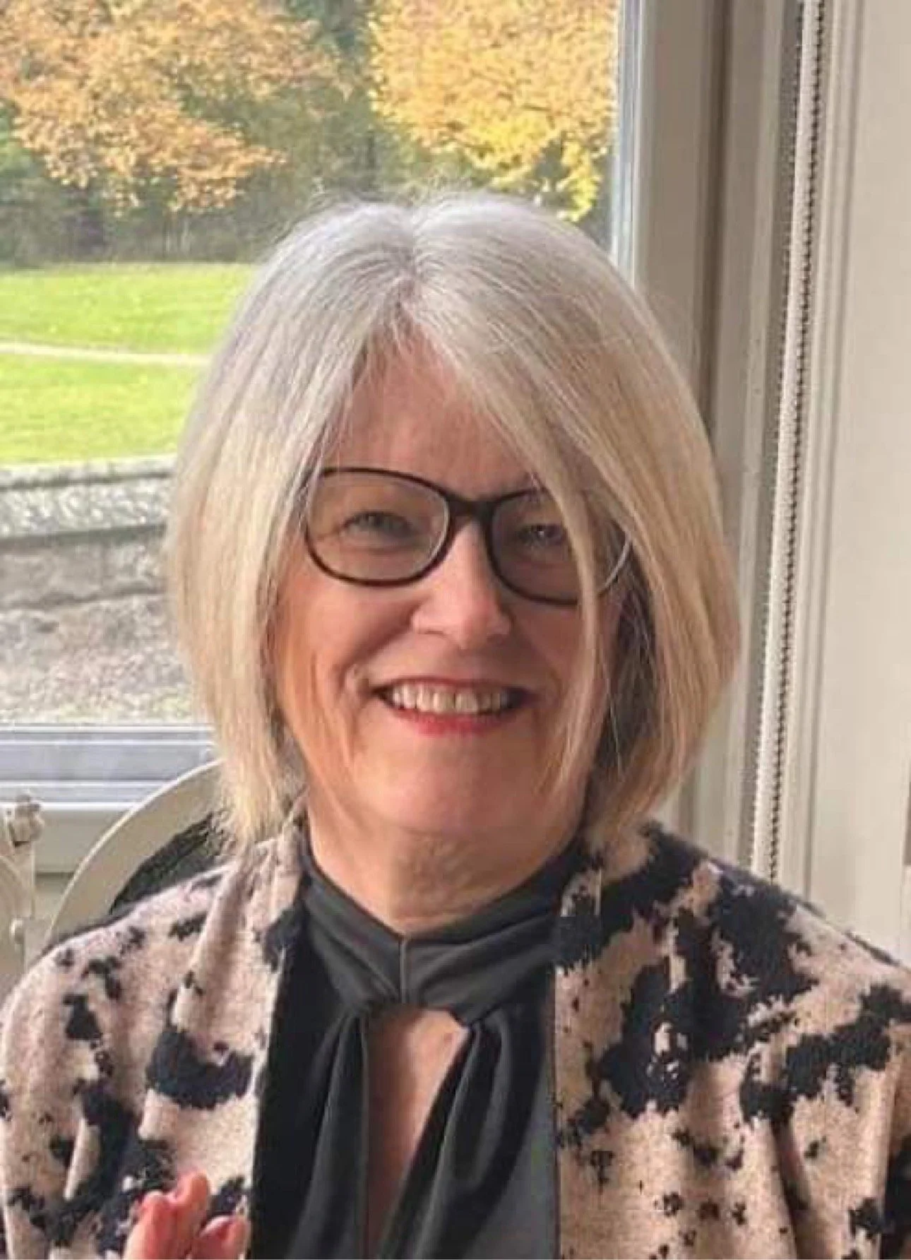Smiling woman with glasses and shoulder-length blonde hair sitting near a window with fall foliage outside.