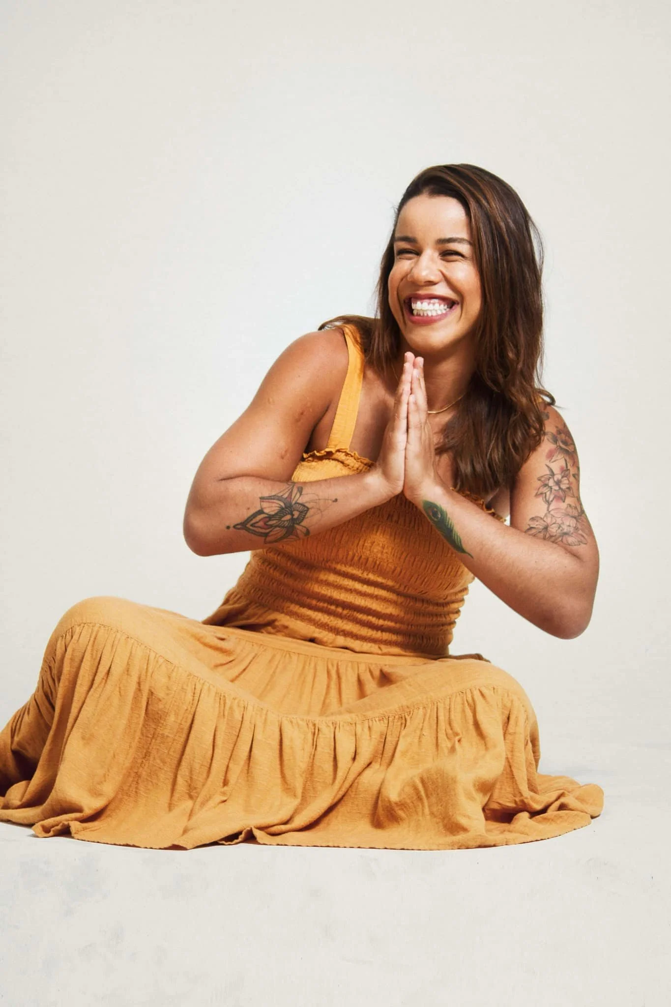 A woman with tattoos on her arms, dressed in a yellow top and skirt, sitting on the floor in a kneeling position with her hands together in a prayer-like gesture, smiling brightly.