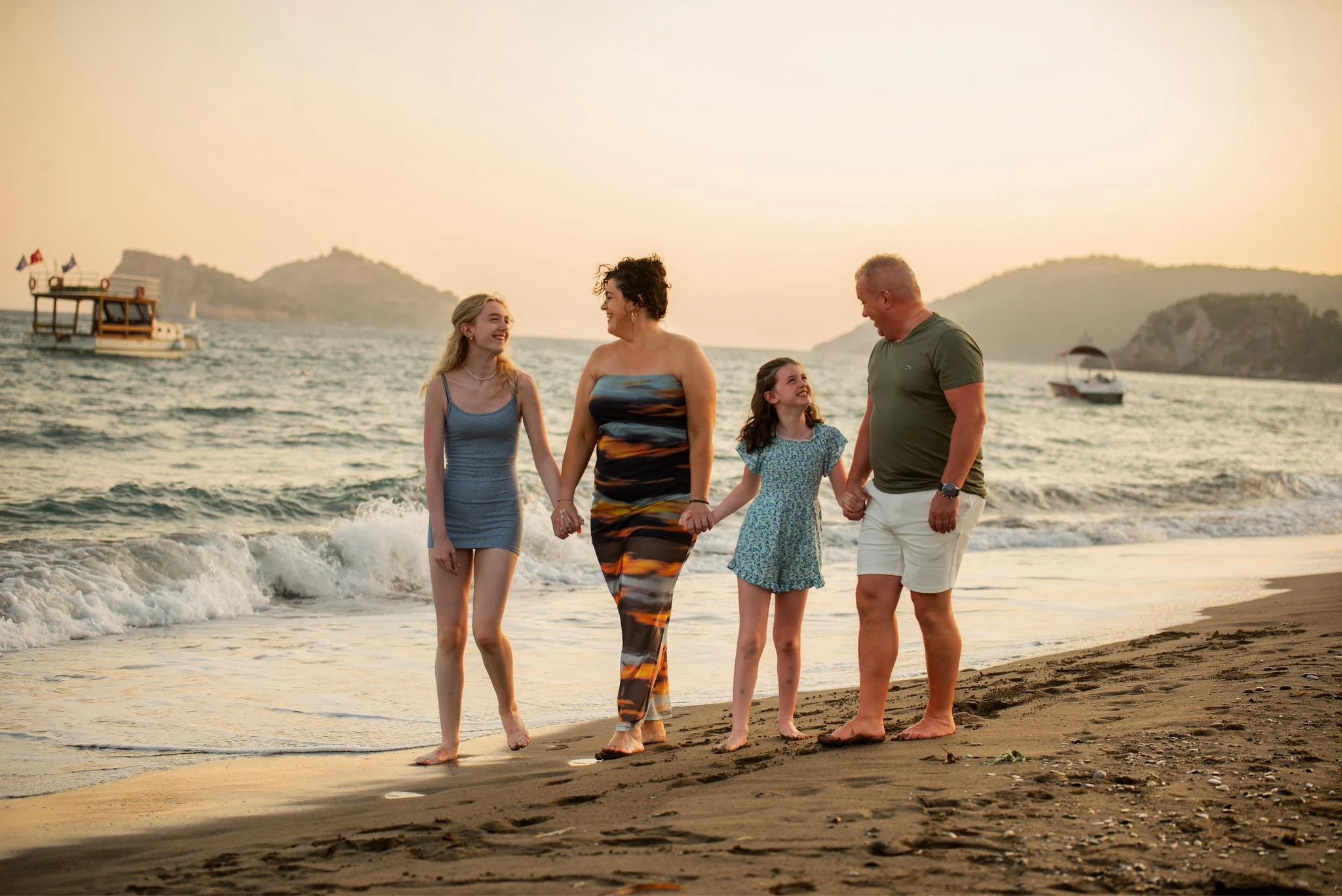 A family of four walking hand-in-hand along the beach at sunset, with boats and islands in the background.
