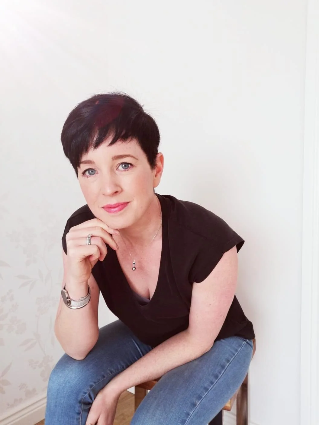 A woman with short black hair, wearing a black top and jeans, sitting on a stool in front of a plain light-colored wall. She is resting her chin on her hand and looking at the camera.