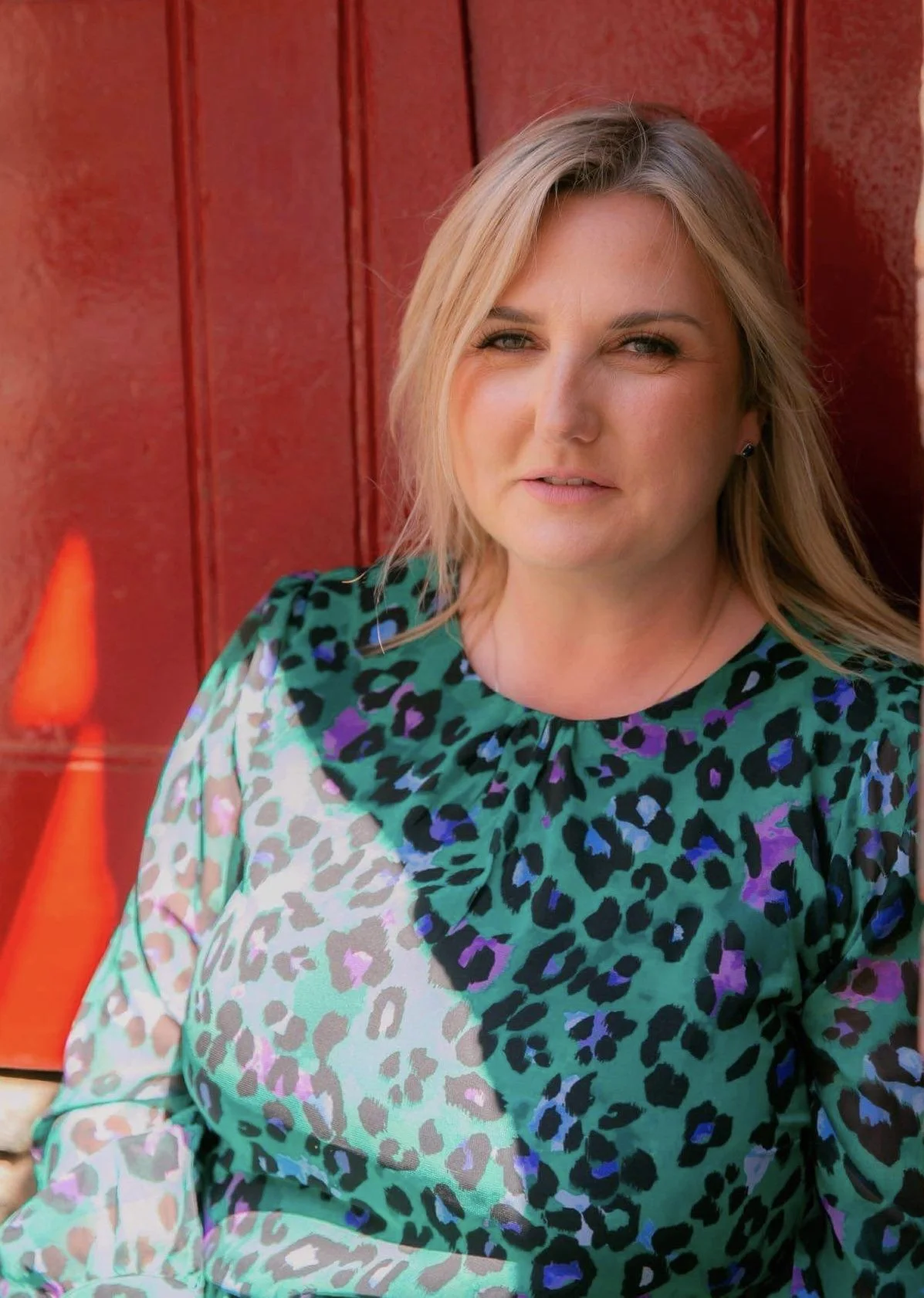 A woman with blonde hair wearing a colorful leopard print blouse, standing against a red wooden background.