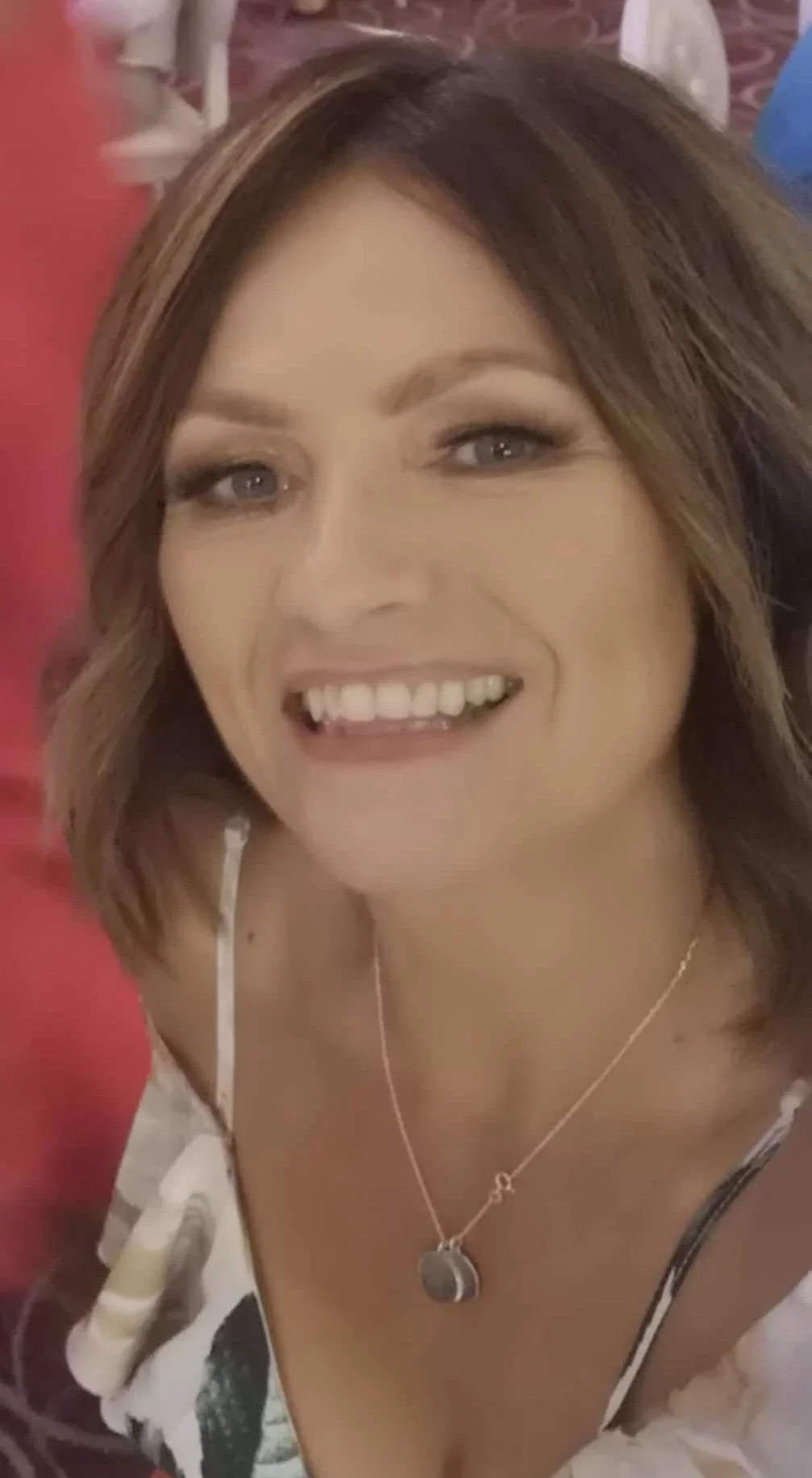 Smiling woman with medium-length brown hair, wearing a necklace with a round pendant, in a setting with red and patterned background.