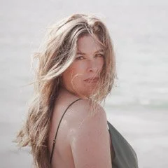 A woman with blonde hair looking over her shoulder at the camera, standing outdoors near water.