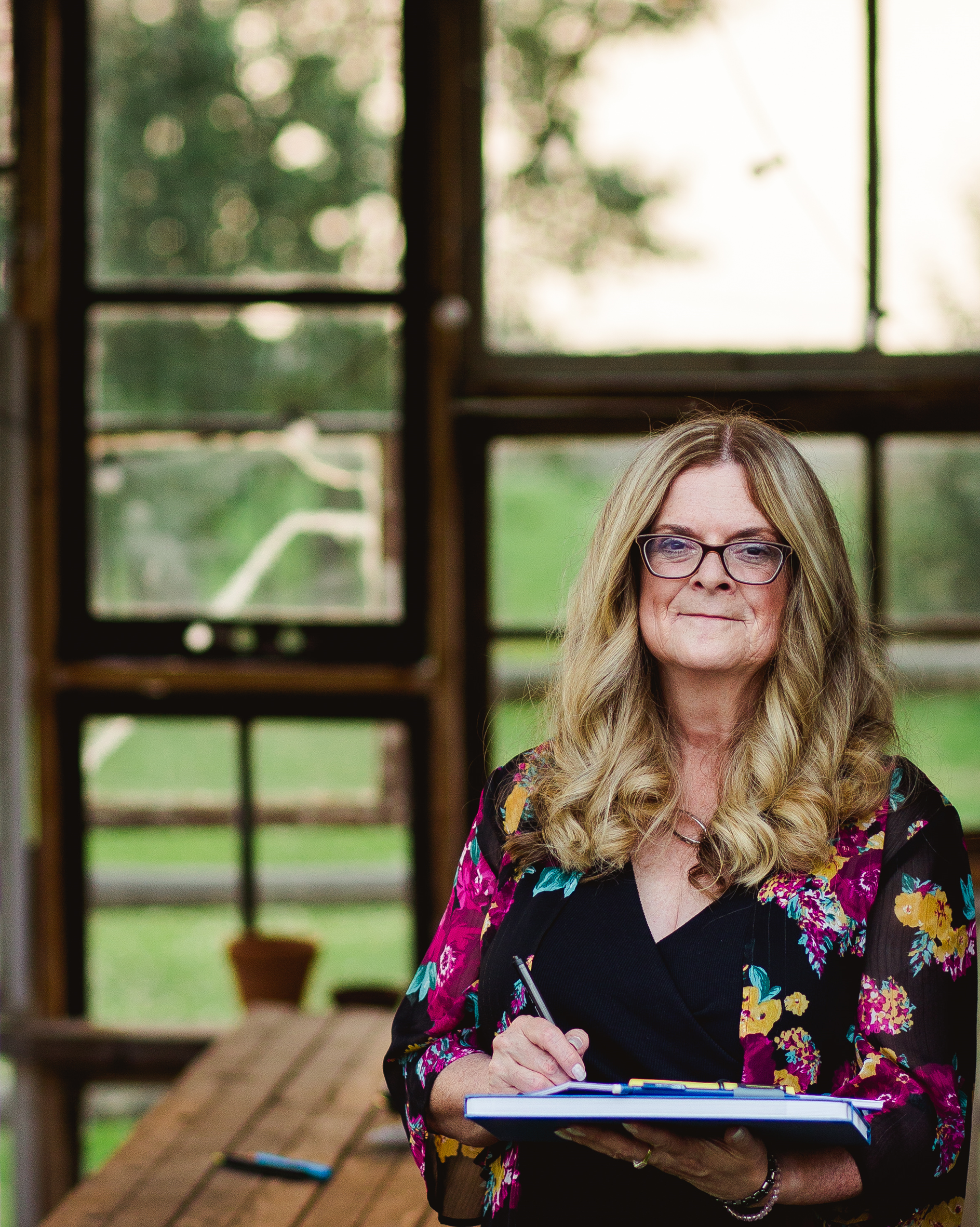 A woman with long blond curly hair and glasses, holding a clipboard and pen, standing in front of windowed walls revealing a green outdoor landscape.
