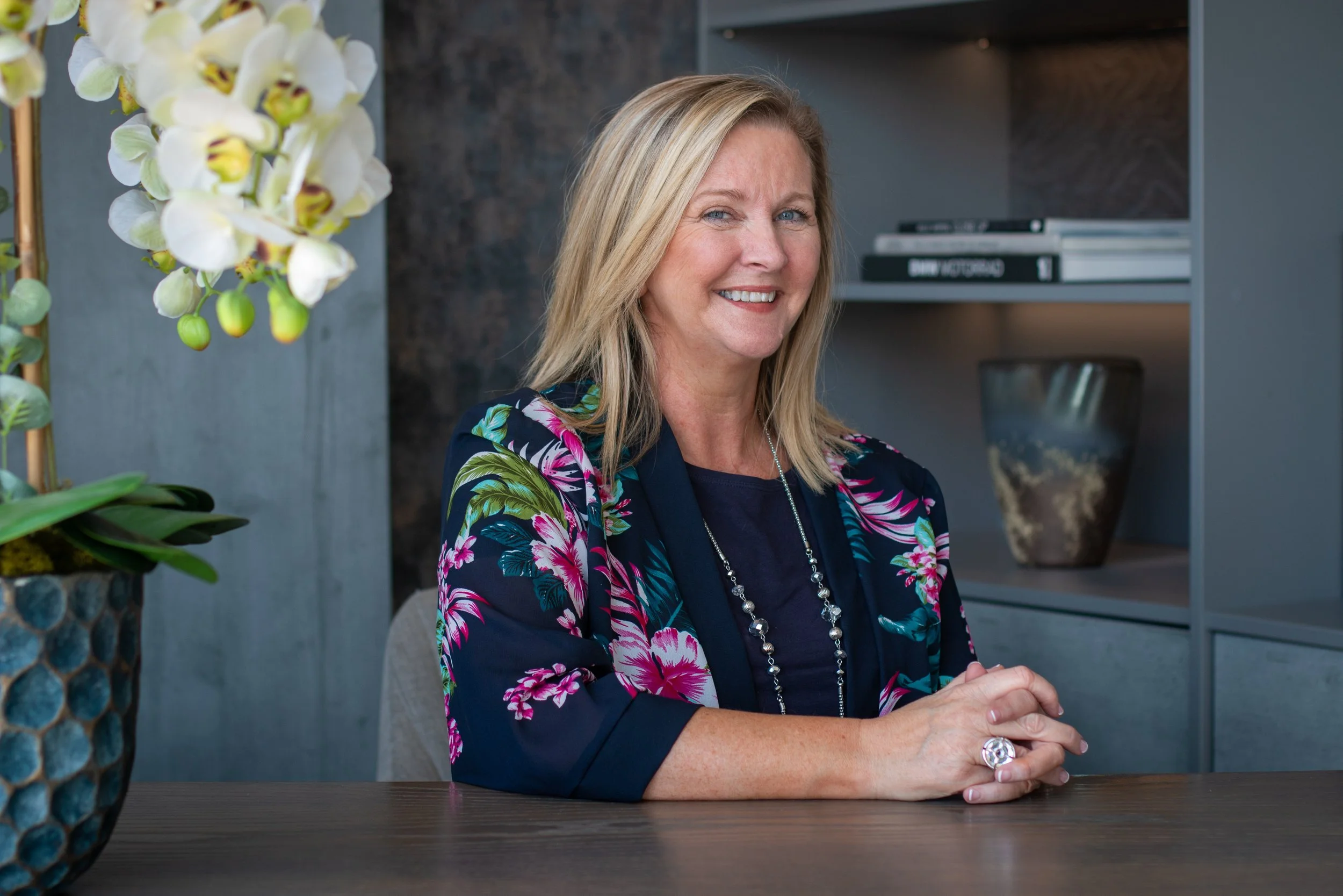 A woman with blonde hair smiling while sitting at a wooden table, decorated with a potted orchid plant.