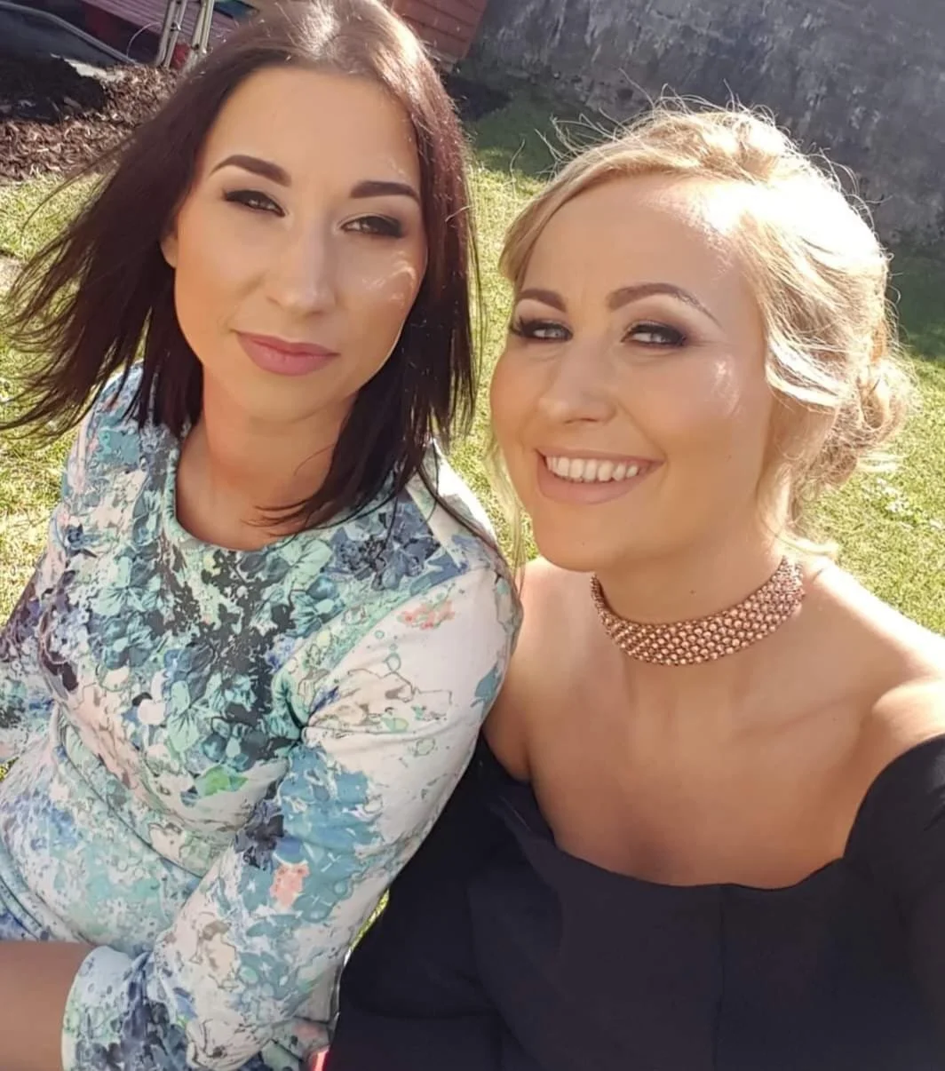 Two women taking a selfie outdoors on a sunny day, both smiling and dressed up, with greenery in the background.