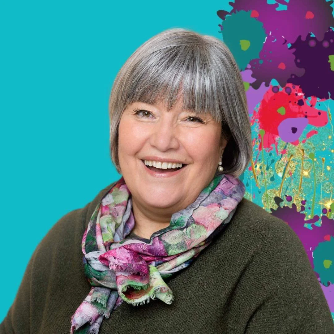 A smiling middle-aged woman with short gray hair wearing a colorful scarf and a dark sweater, with a vibrant graffiti-inspired background.