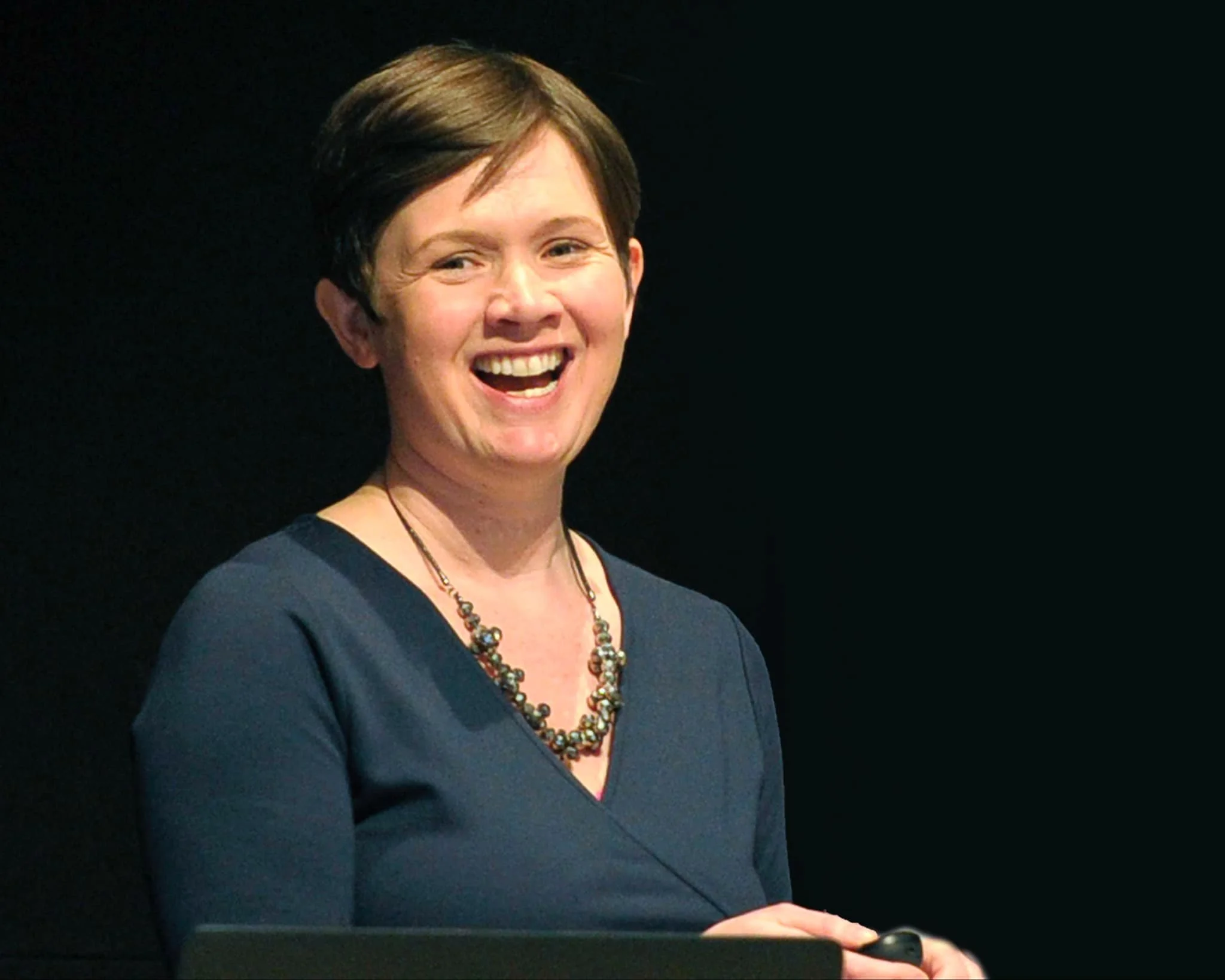 A woman with short dark hair smiling and wearing a dark blue top and patterned necklace, standing in front of a black background.