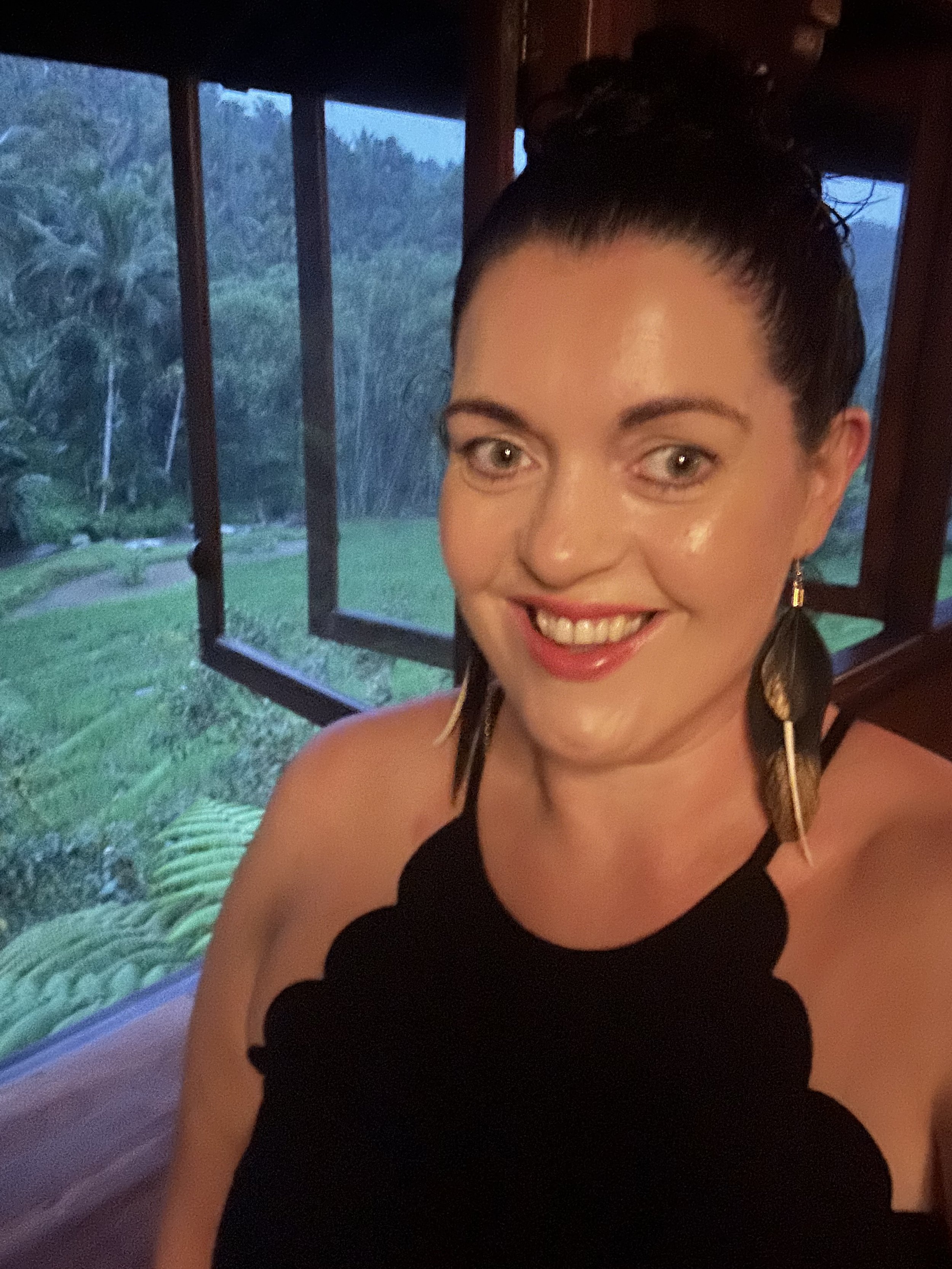 A woman with dark hair in a bun, wearing a black dress and large feather earrings, smiling indoors with open windows revealing a lush green outdoor landscape.