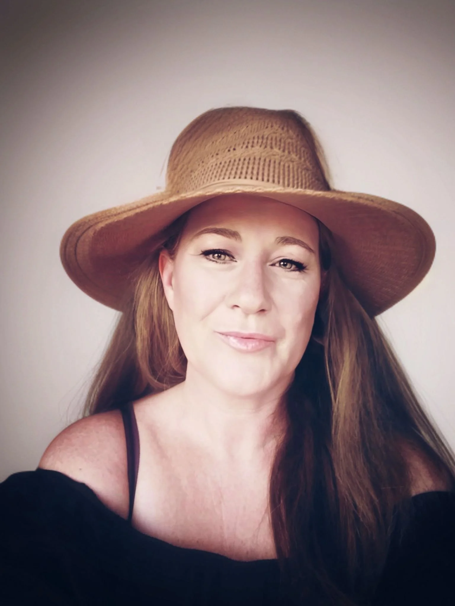 A woman with long reddish-brown hair wearing a wide-brimmed straw hat and a black off-the-shoulder top.