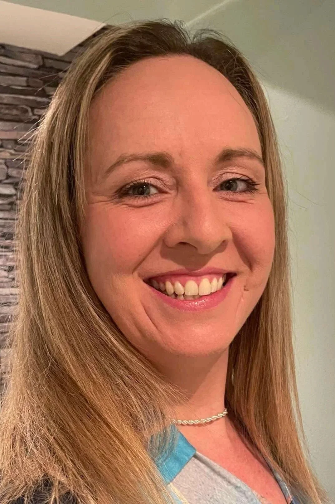 Close-up of a smiling woman with long blonde hair, wearing a necklace and a top with blue and gray colors.