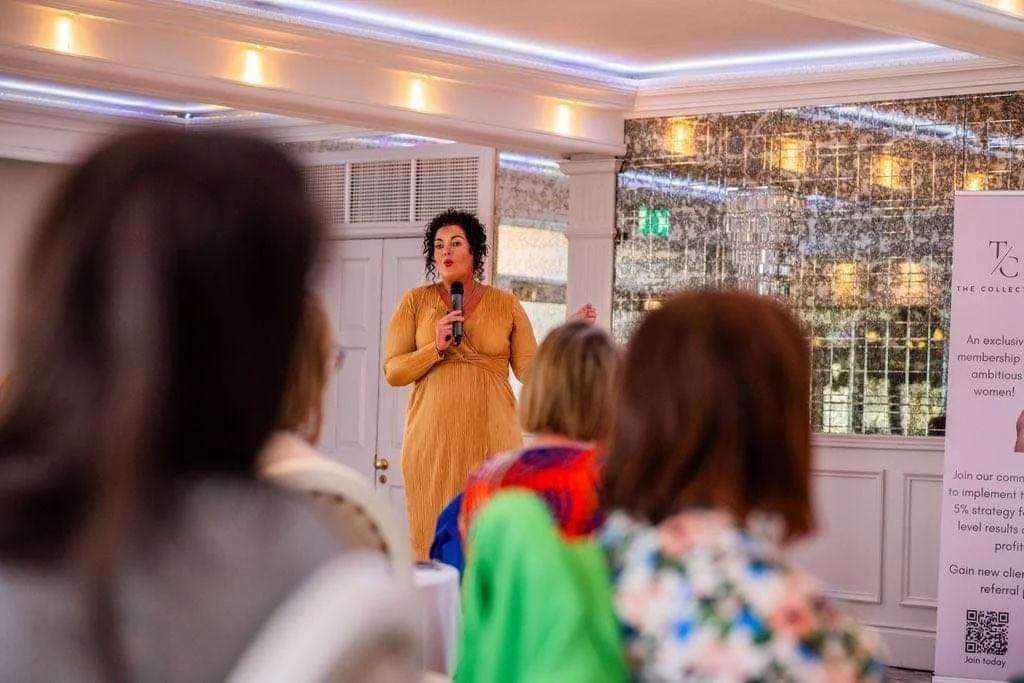A woman in a yellow dress giving a presentation or speech with a microphone at a formal indoor event. Several audience members are seated, listening attentively.