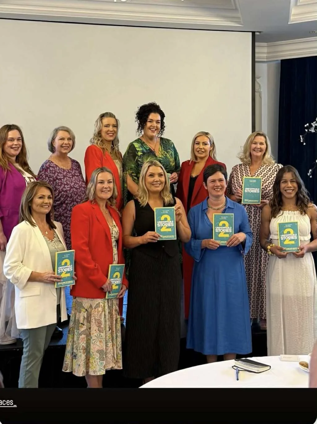 Group of women at an event, holding books titled 'Story 2' with a distinctive yellow and blue cover, standing in front of a blank presentation screen.
