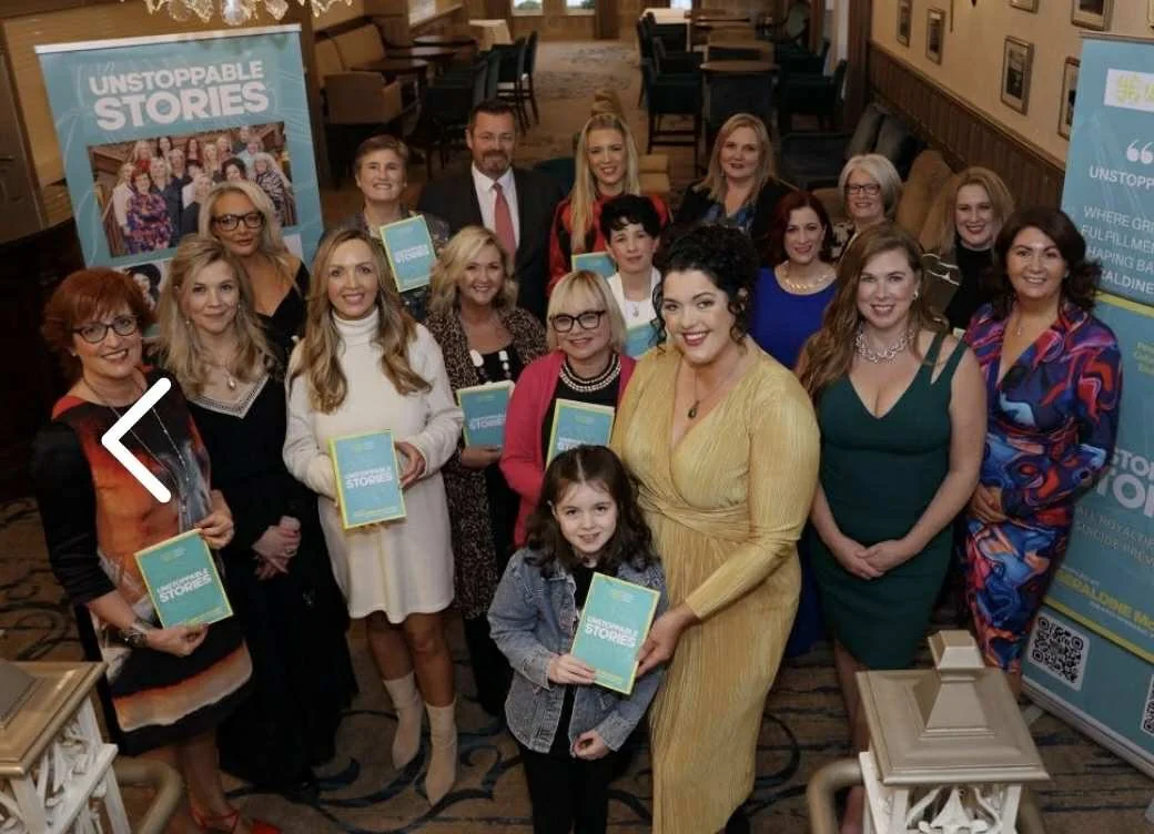 Group of women and one girl attending an event, holding copies of a book titled "Unstoppable Stories," inside a room with tables and framed pictures on the walls.
