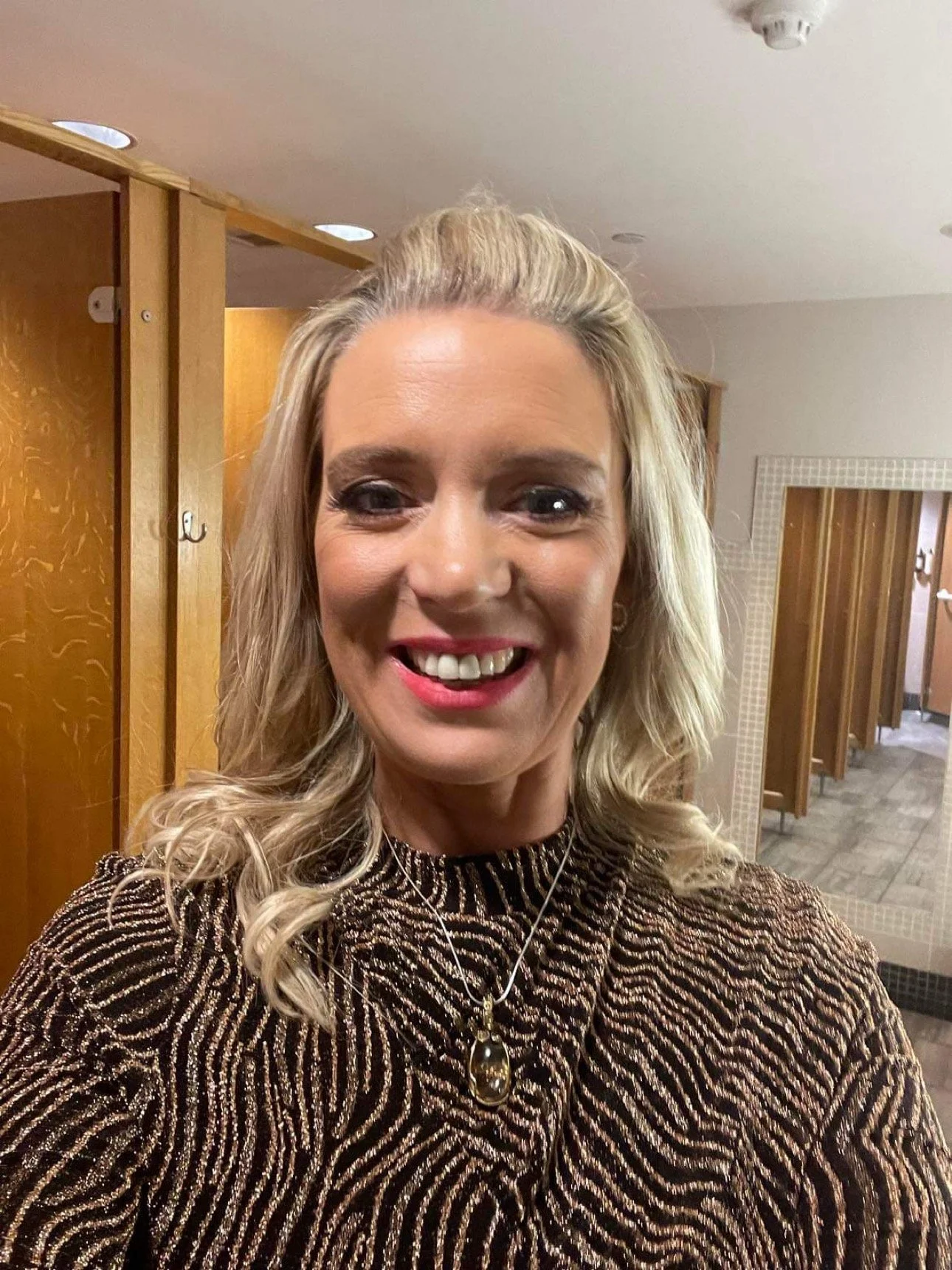 A smiling woman taking a selfie in a room with wooden lockers, wearing a patterned black and gold top and a necklace.
