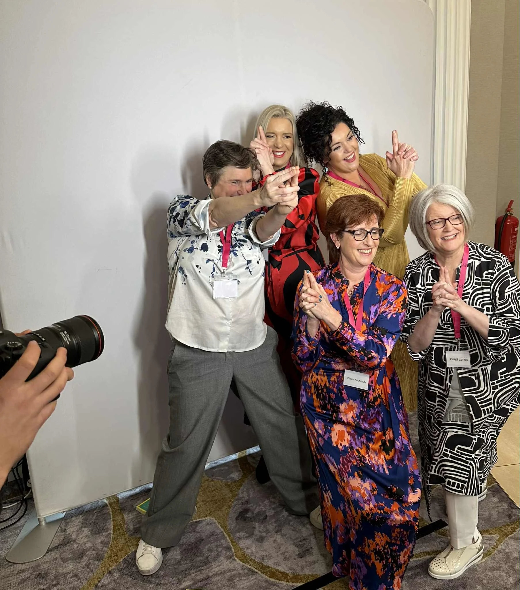 Five women smiling and pointing at a camera, standing against a plain white wall at an indoor event, with 2 women in the front and 3 in the back, all wearing colorful and patterned clothing.