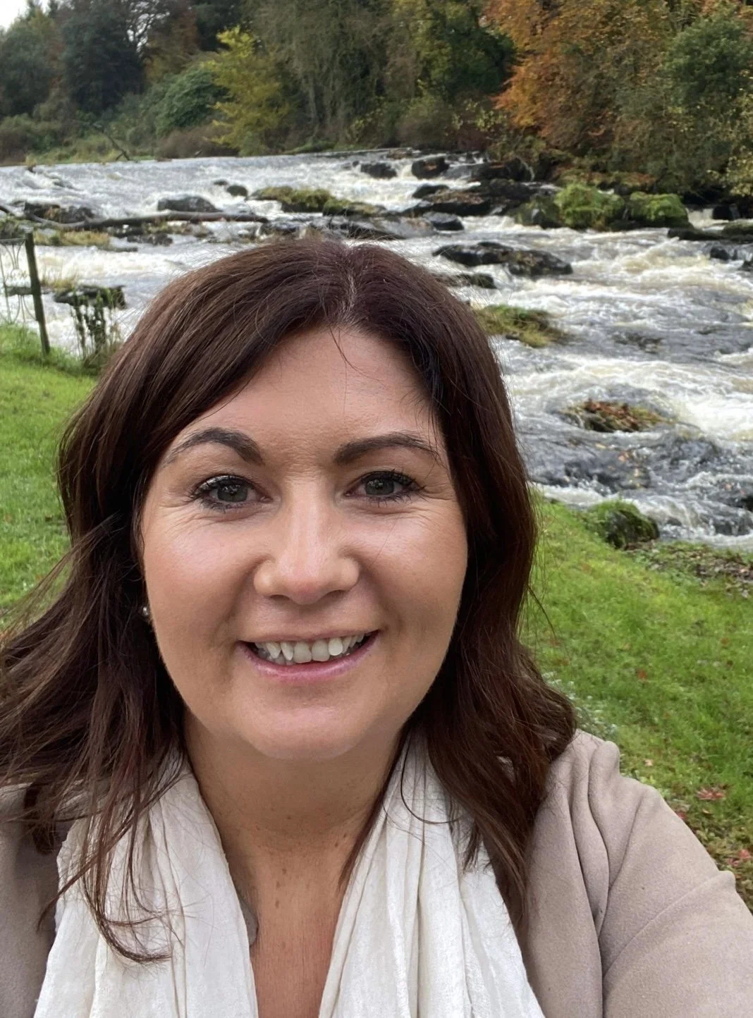 A woman smiling outdoors with a river and trees in the background.