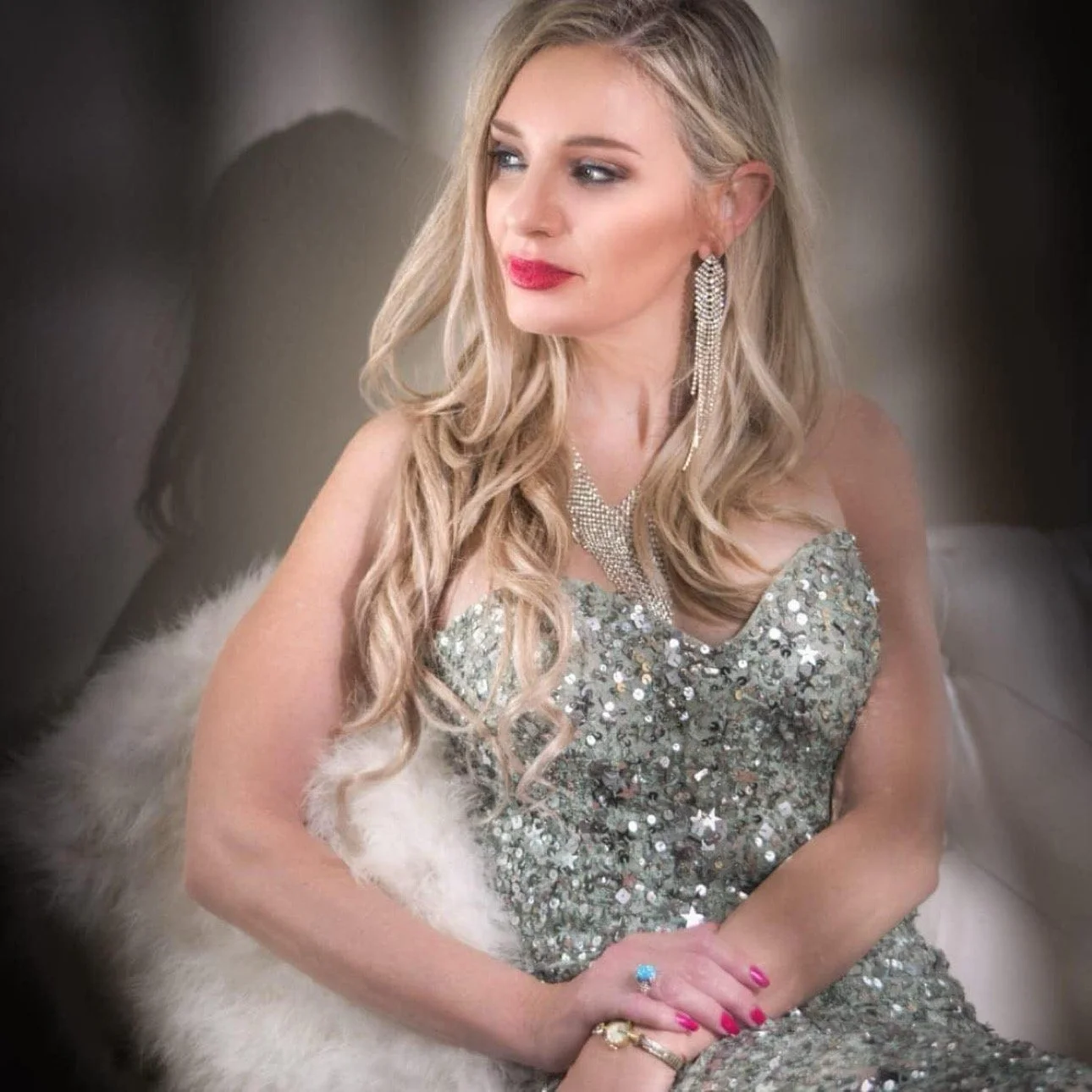 A woman with long blonde wavy hair, wearing a sparkly silver dress and jewelry, sitting on a furry white surface.