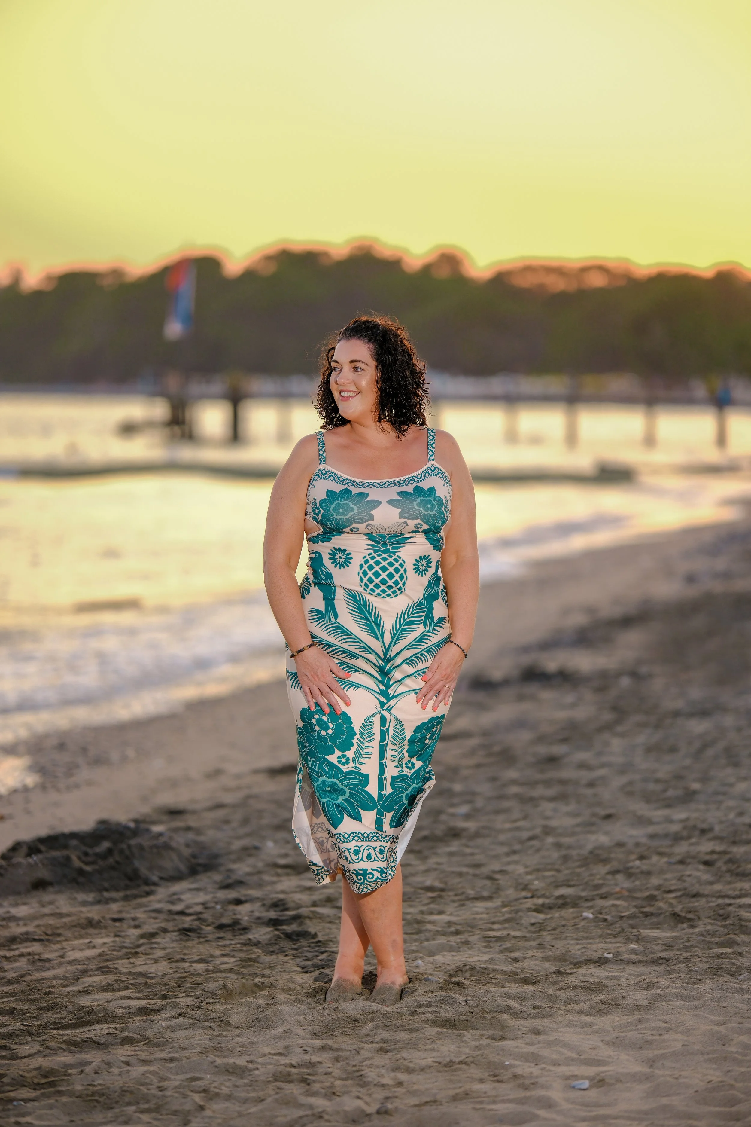 A woman in a patterned maxi dress walking barefoot on the beach at sunset, smiling and looking to the side.