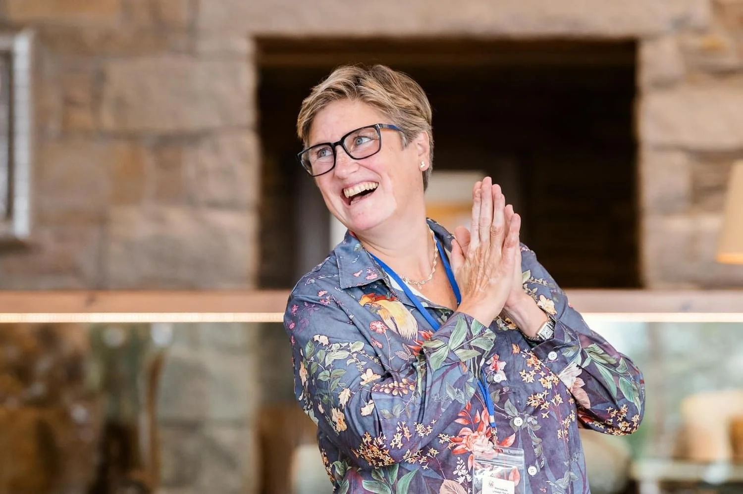 A woman with short hair, glasses, and a floral blouse, smiling and clasping her hands together in front of a brick fireplace.