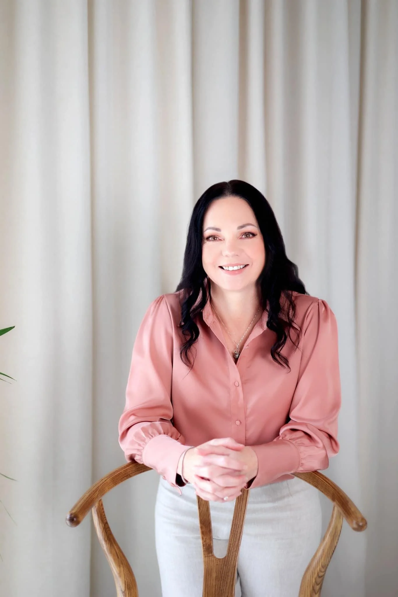 A woman with black hair smiling, wearing a pink blouse and light-colored pants, sitting behind a wooden chair in front of a cream-colored curtain.