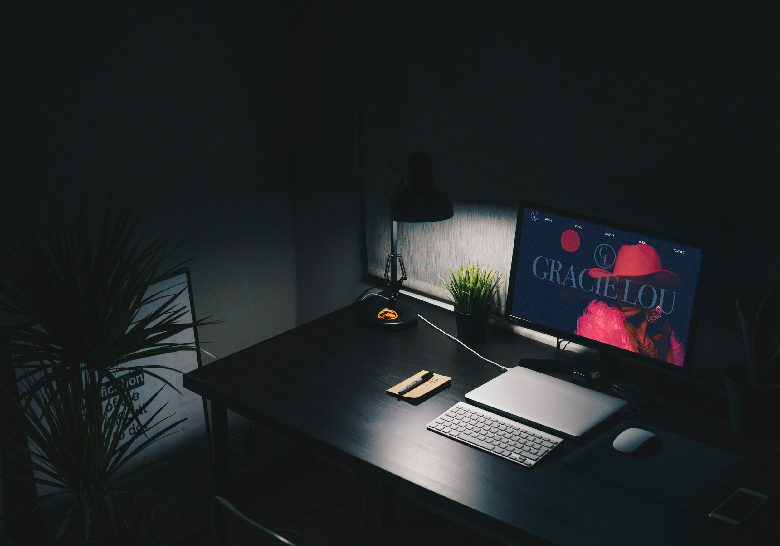 Dark workspace with a computer monitor displaying a fashion website, a closed laptop, keyboard, mouse, desk lamp, potted plant, notepad with pen, and a large plant on the side.
