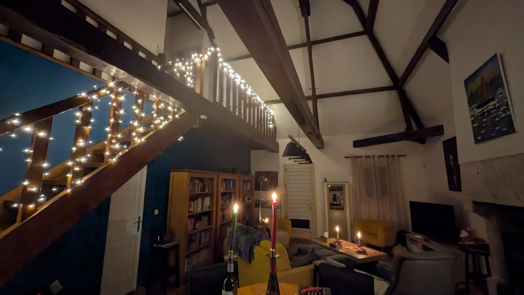 Living room with dim lighting, decorated with string lights on the staircase, candles on tables, bookshelf, armchairs, and artwork on the walls.